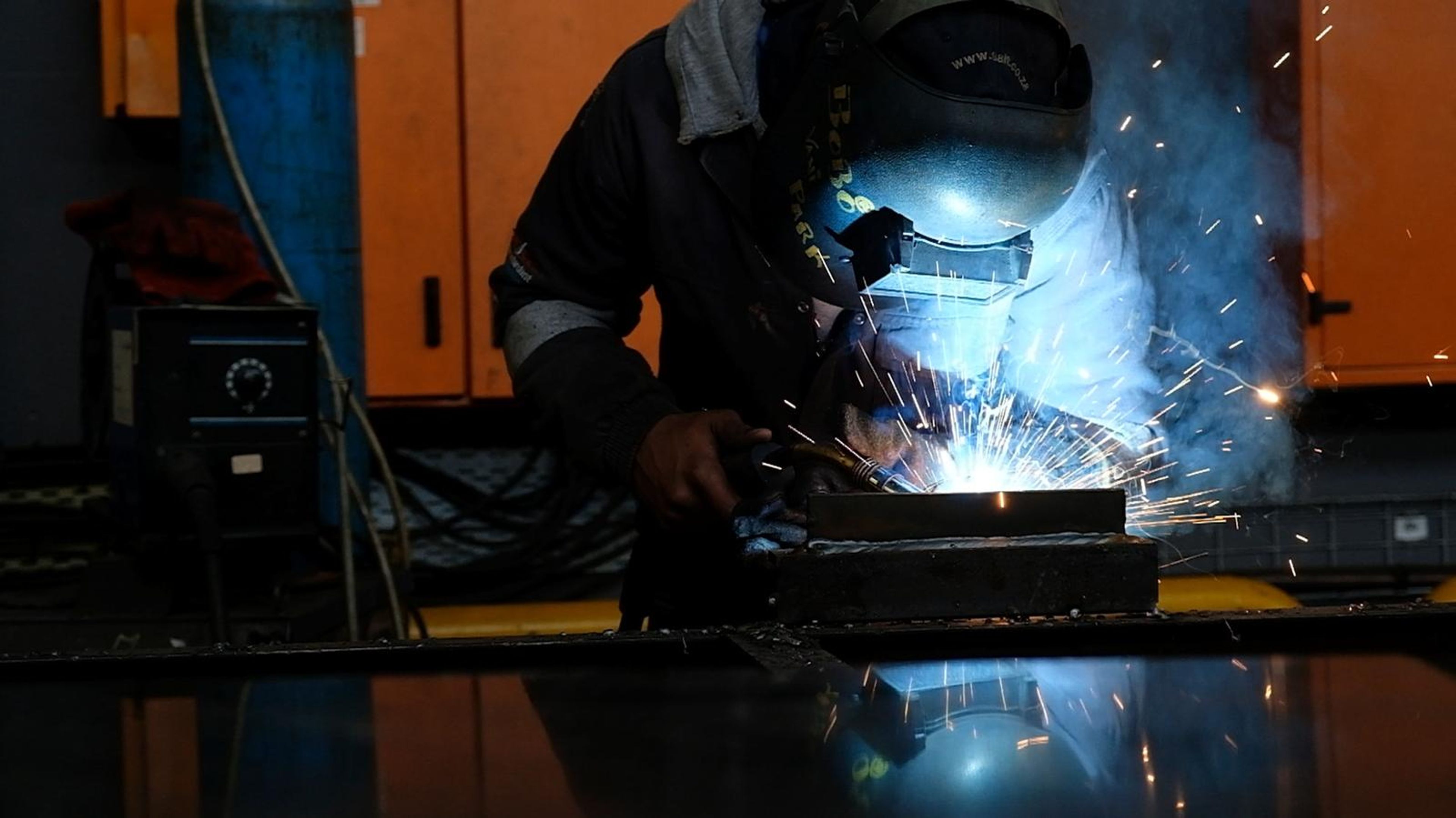 Welder in a mask at a steel bar in front of orange cabinets, arc glowing and sparks flying