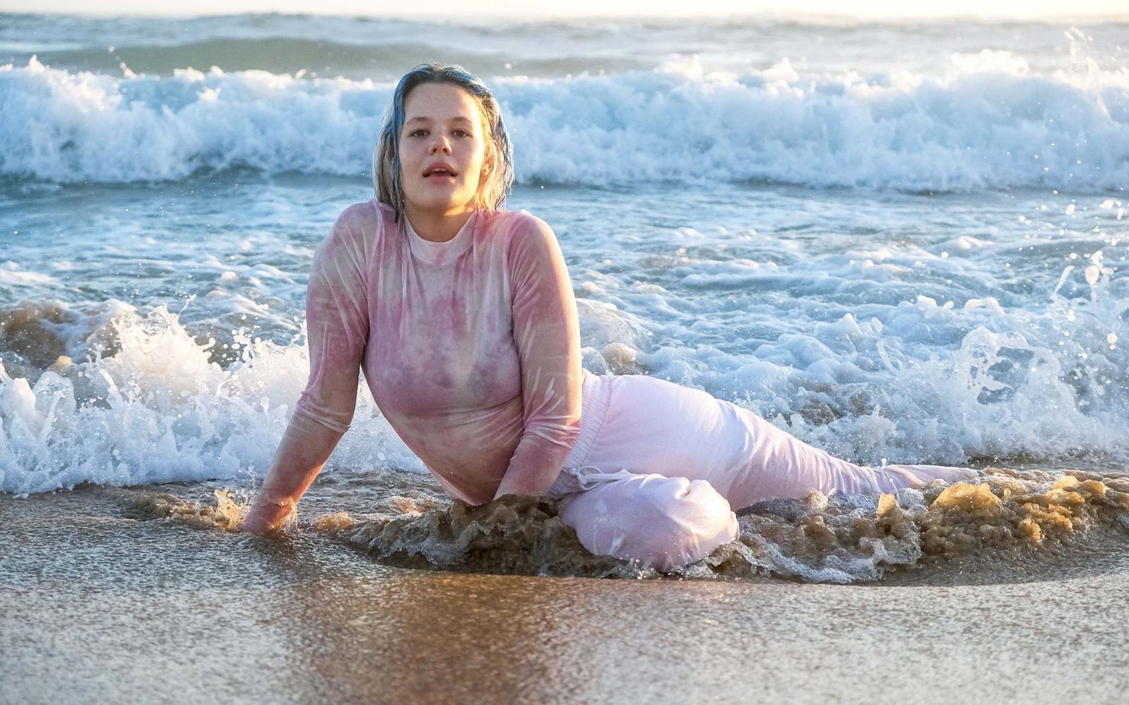 Woman reclining in the shallows with one arm braced, wet pink top, parted lips, whitewater behind her