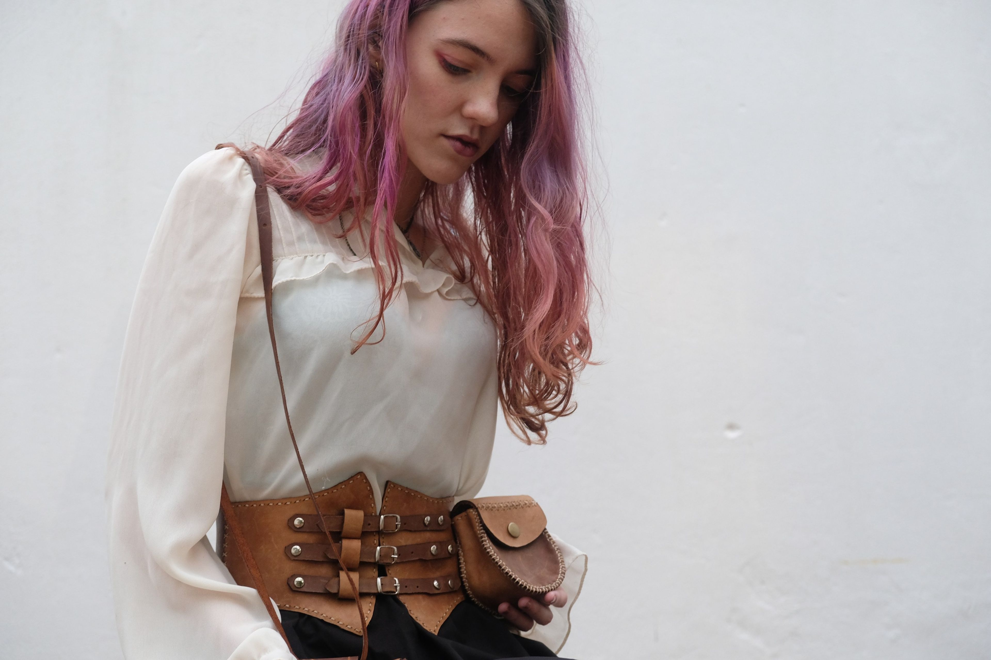 Woman with pink hair in a cream blouse wearing a tan leather corset belt with a pouch