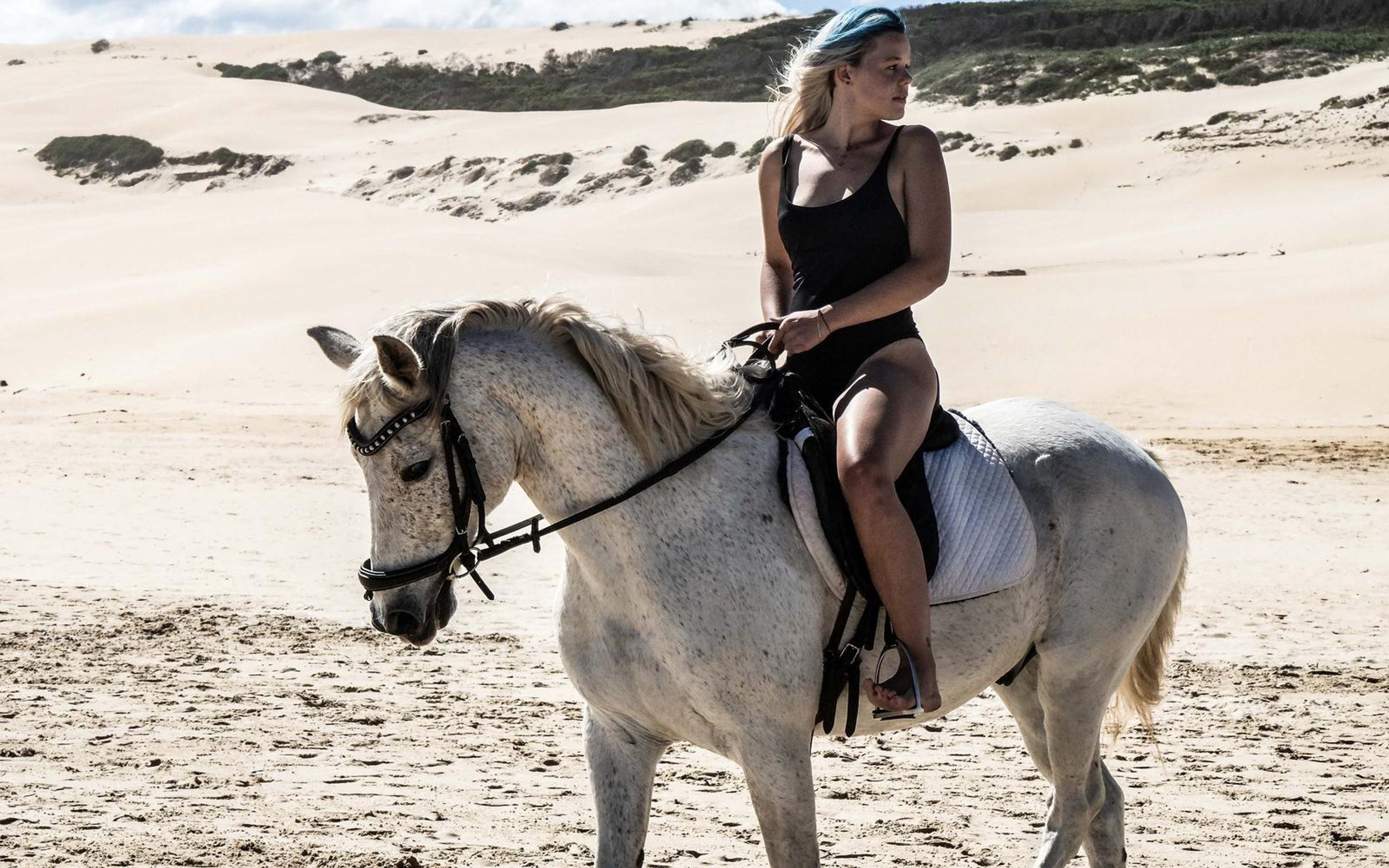 Rider seated on a white horse on pale sand, looking toward the camera, dunes behind