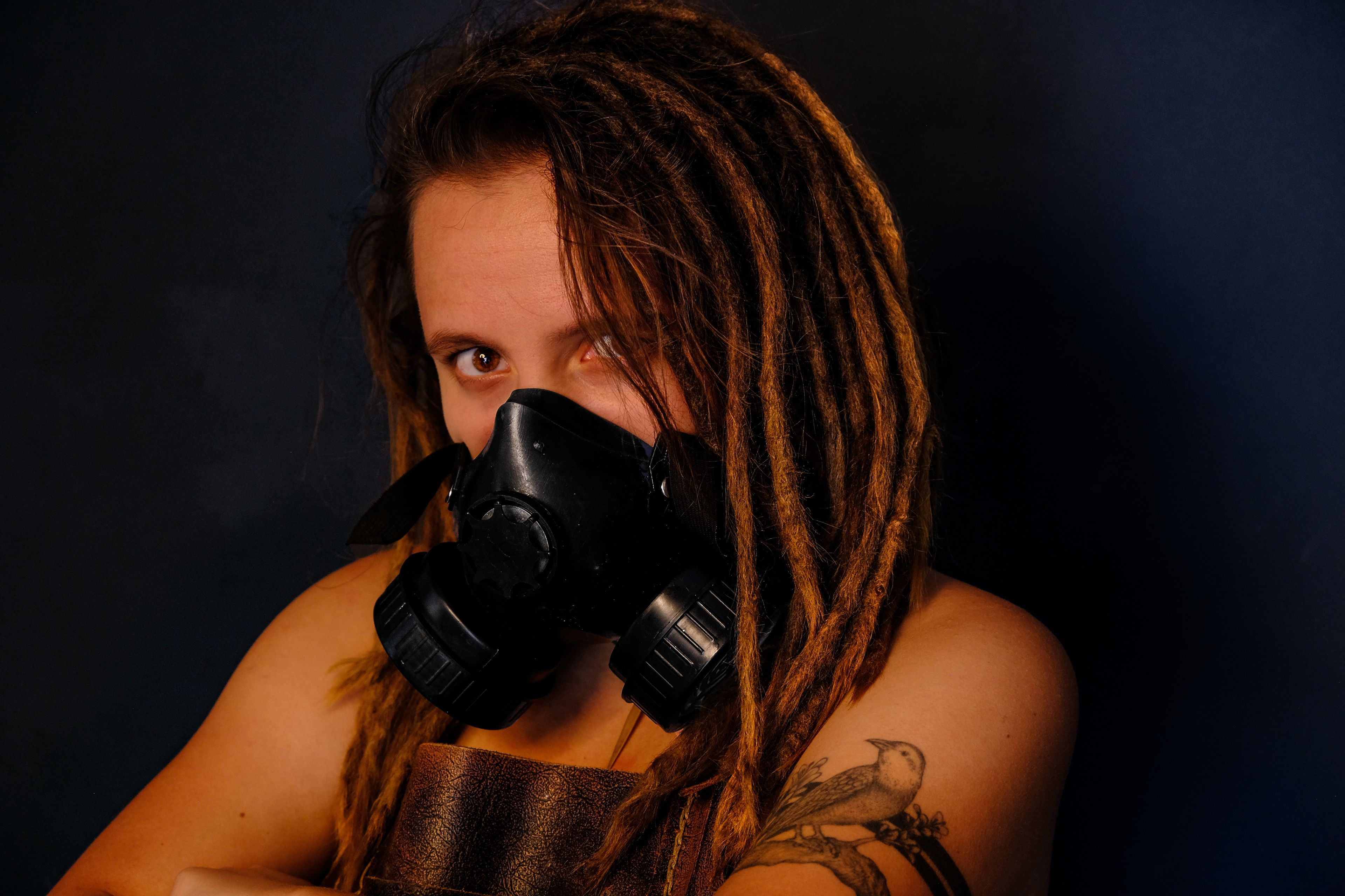 Warm-toned portrait, subject wearing a gas mask, dreadlocks framing the face, bird tattoo visible on the shoulder