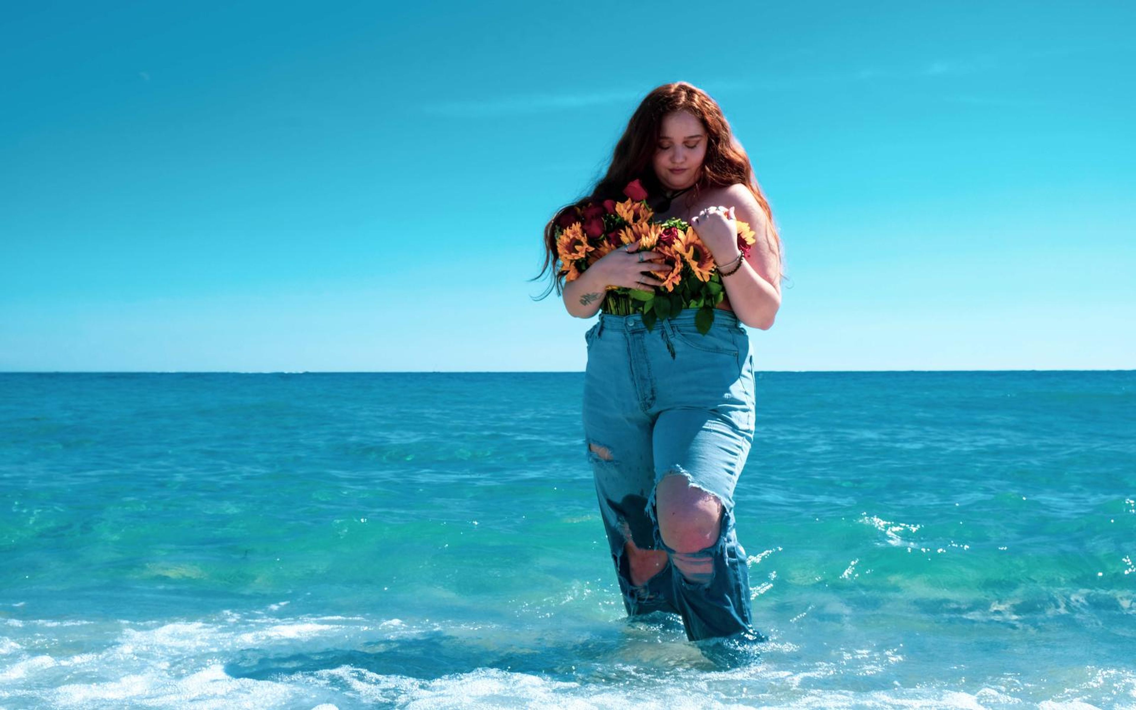 Model in a deep cyan toned frame, standing in the water with bouquet, looking down, wide sky