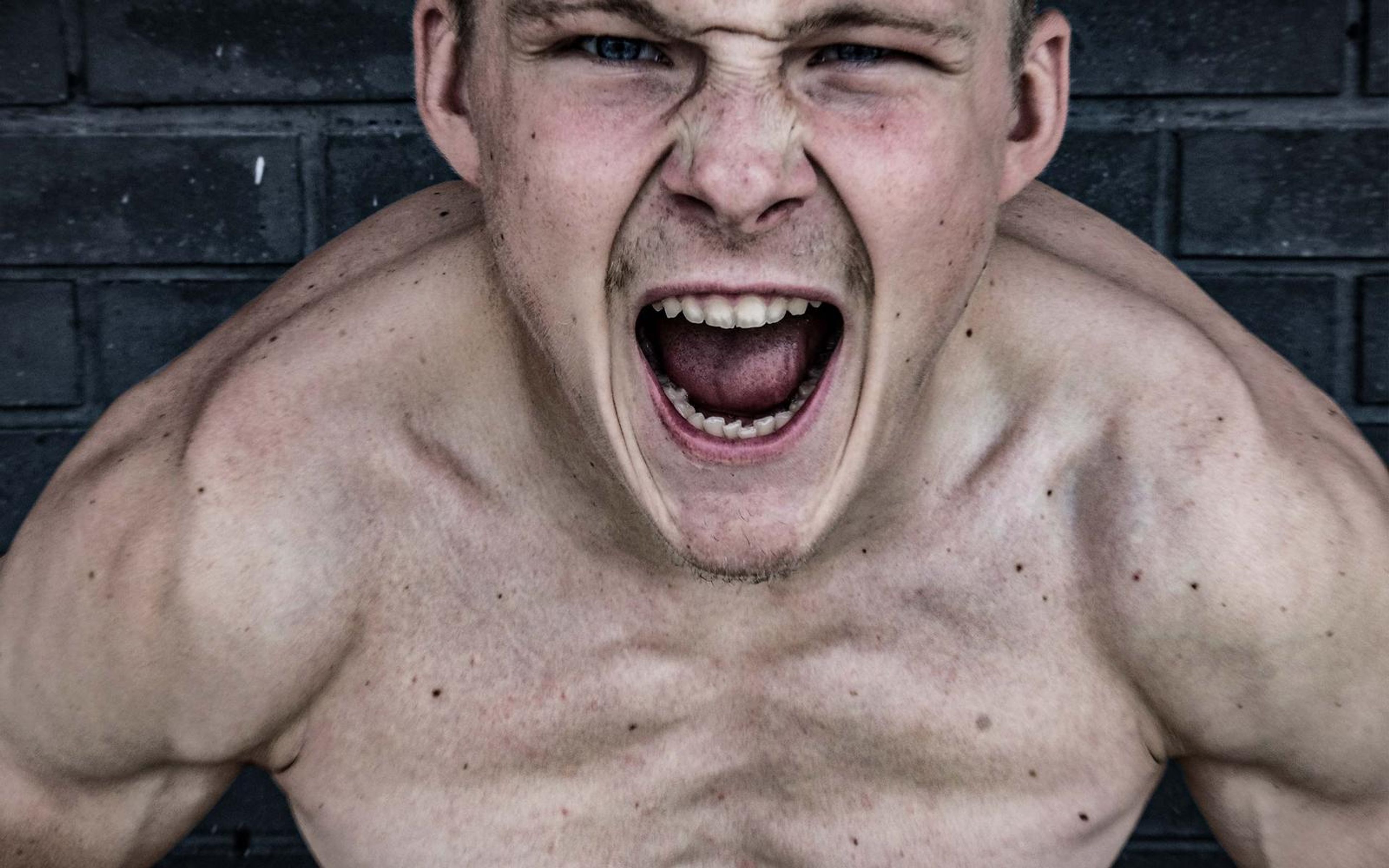 Man shouting at camera, shoulders hunched forward, shirtless against a dark brick wall