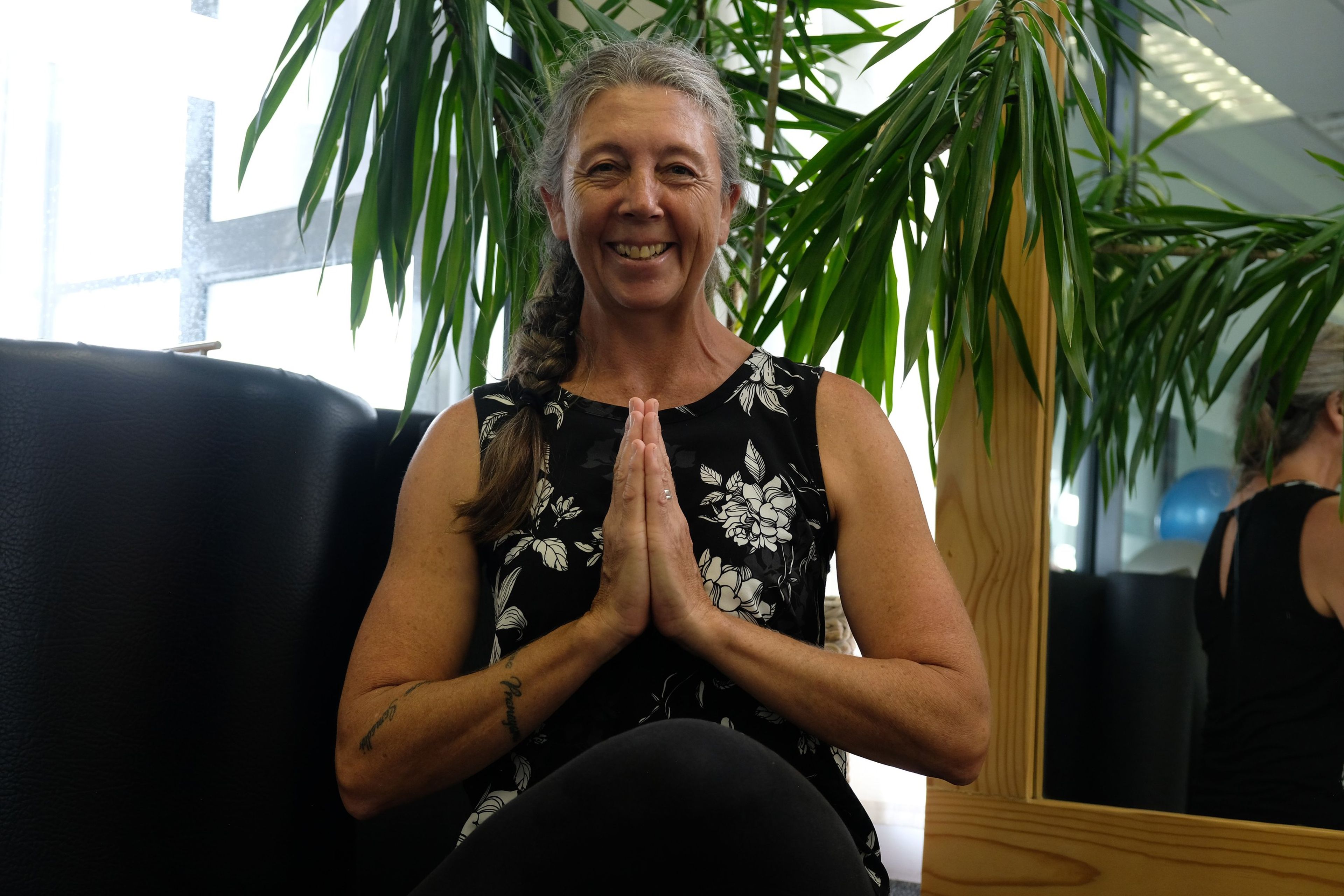 Seated instructor in a floral top with hands in prayer position, a plant and mirror behind her