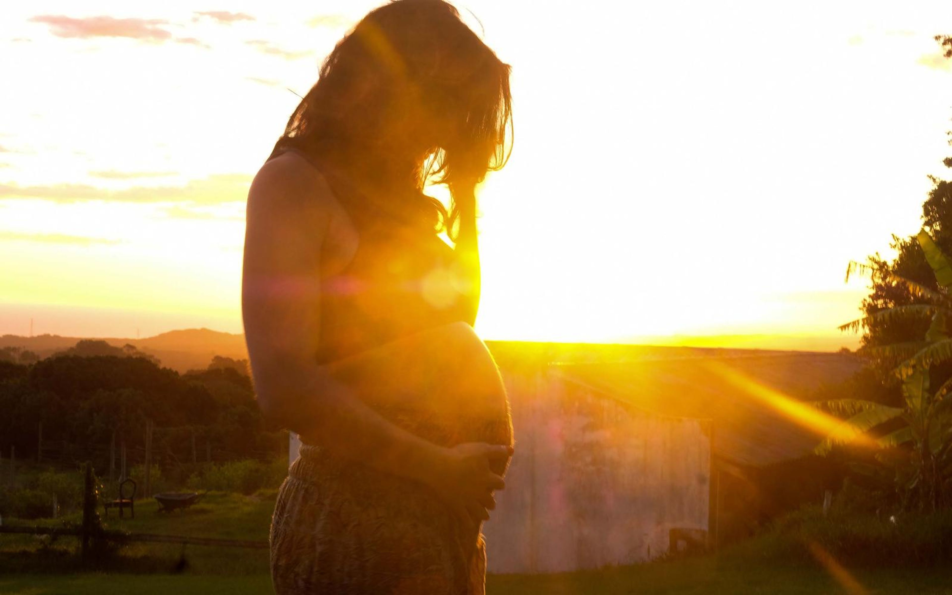 Backlit pregnant woman with long hair falling forward, hand at her belly, golden flare, trees behind