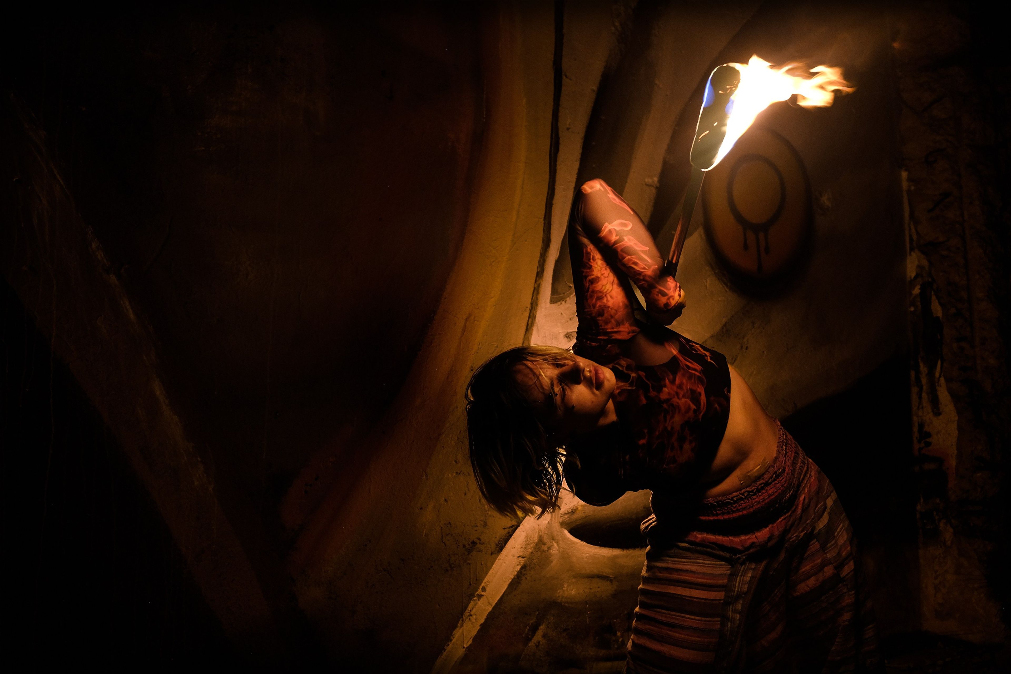 Performer in patterned trousers holding a fire staff horizontally in front of a mural with an orange circle emblem