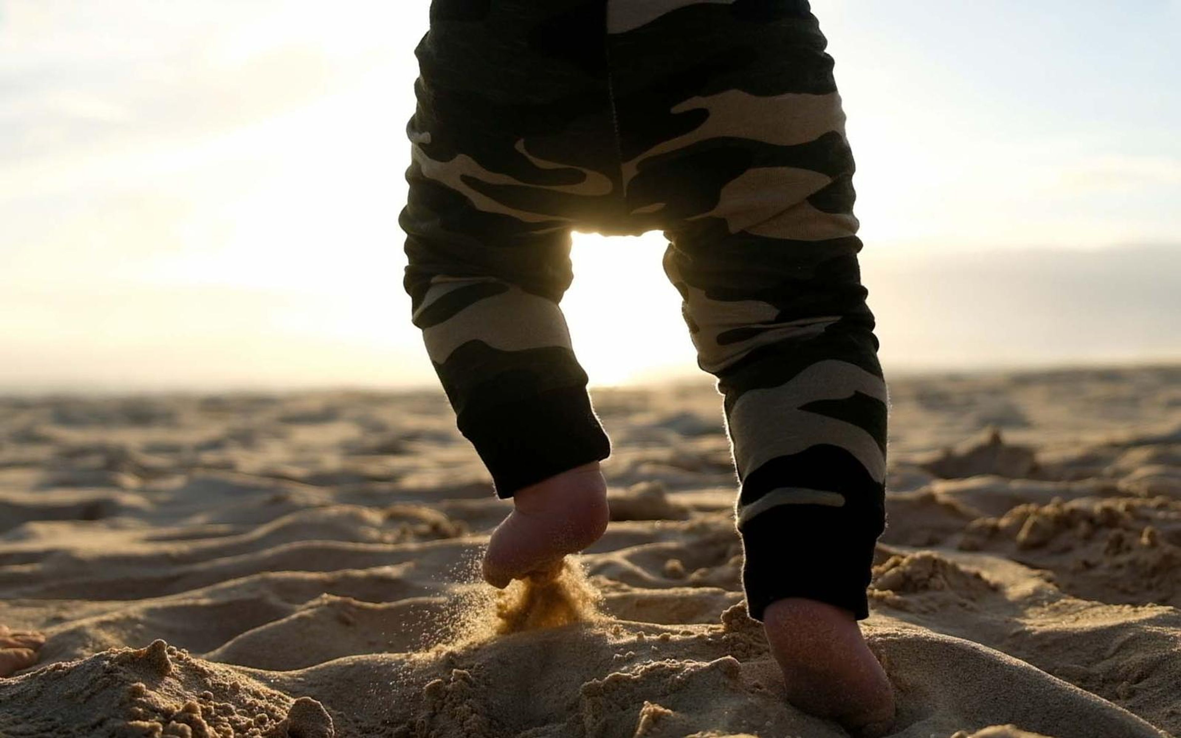 Low view of a toddler in camouflage trousers walking barefoot on sunlit sand with the sun flaring between the legs