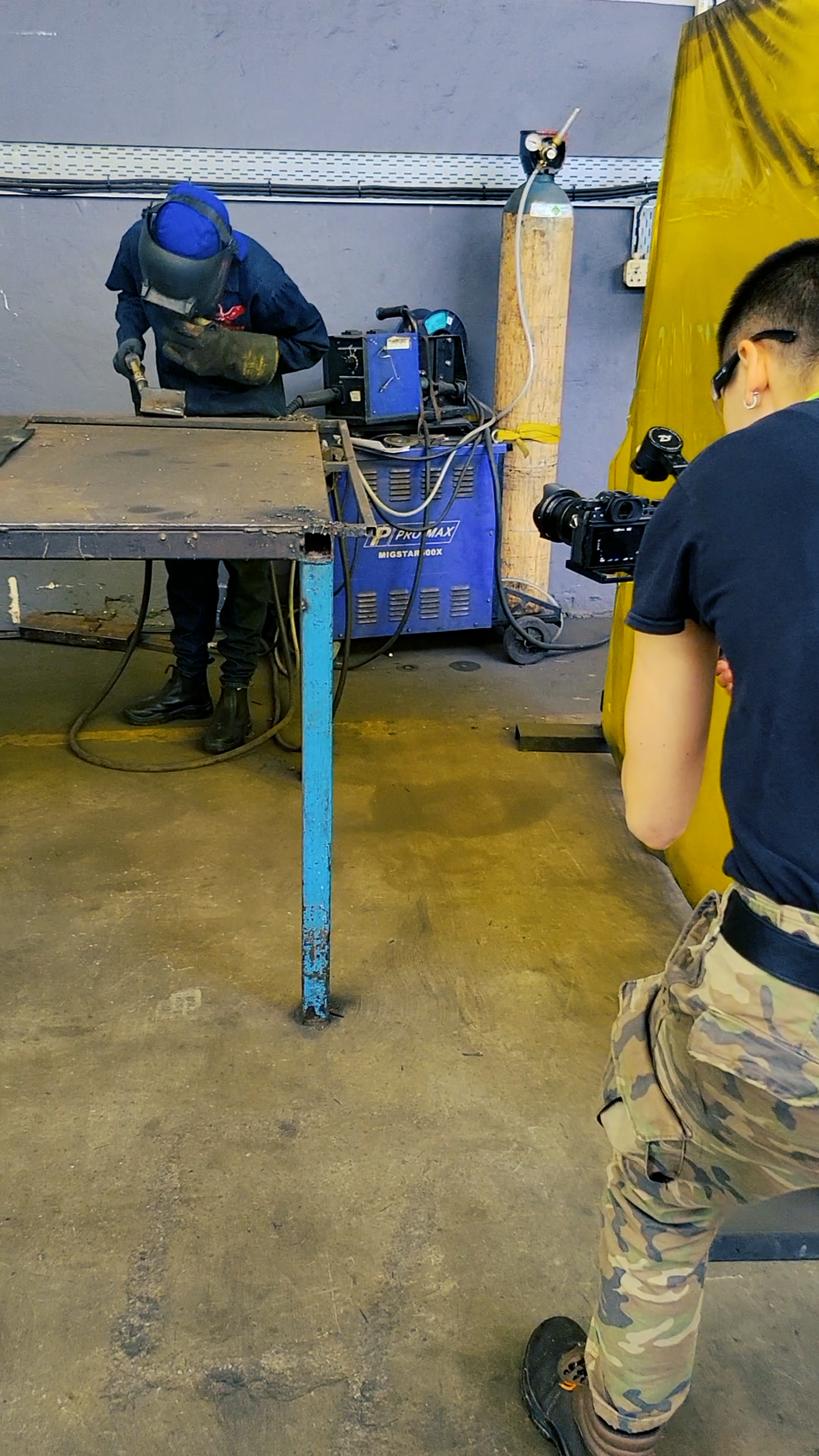 Behind the scenes in a workshop, a camera operator filming a welder beside a yellow weld screen