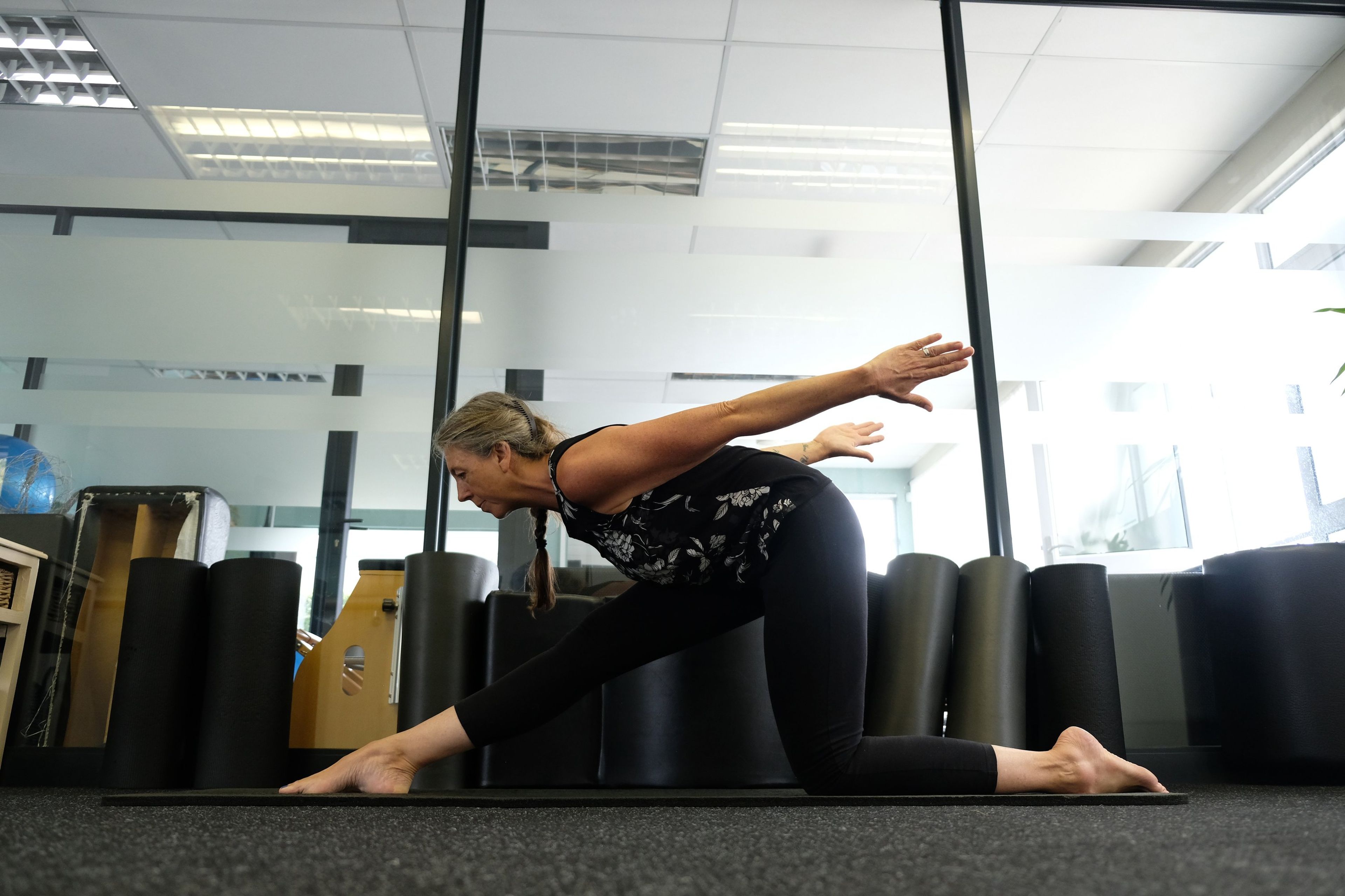Instructor in a deep lunge with arms extended, foam rollers and studio equipment behind her