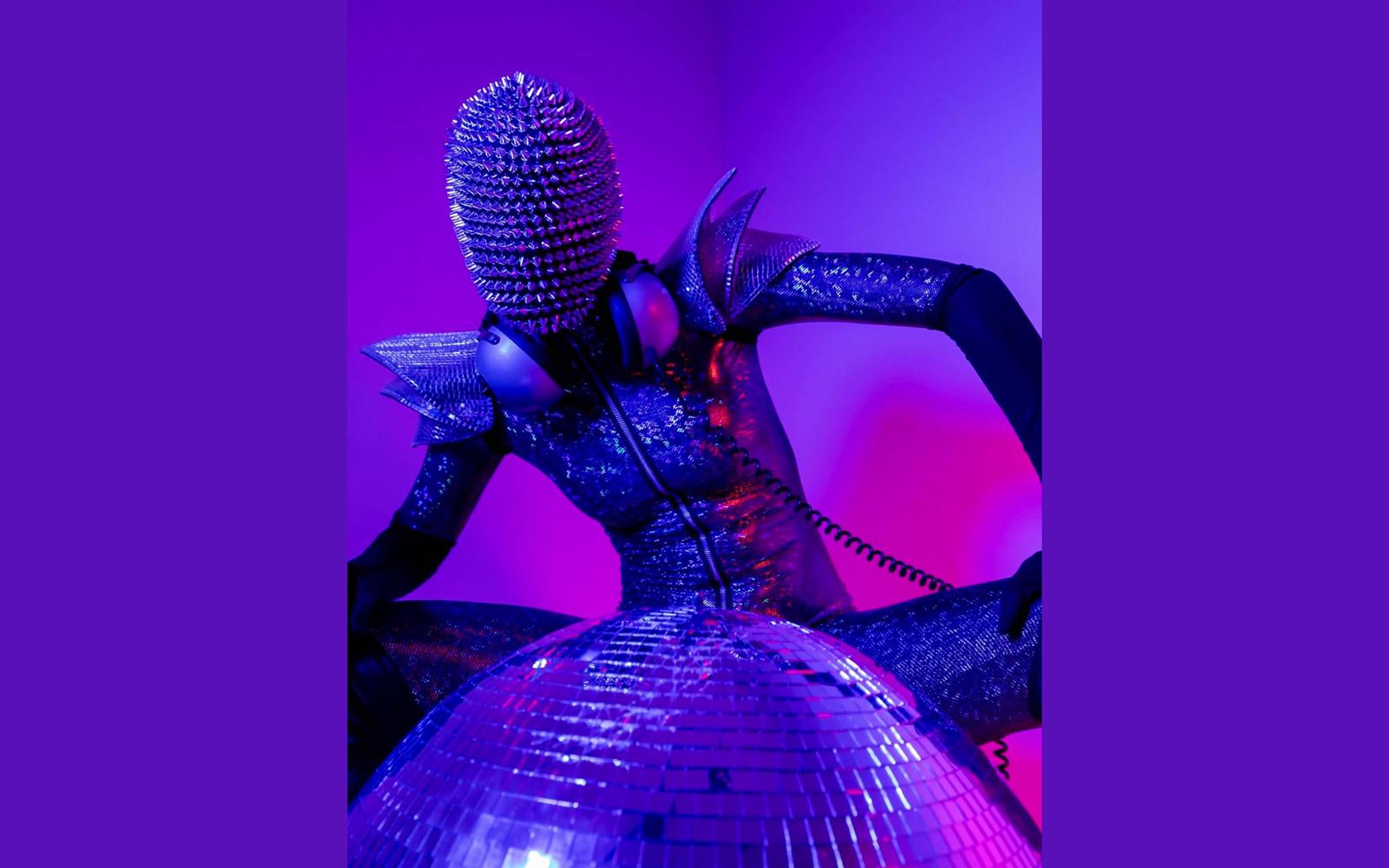 Performer in a spiked mirrored mask and sequinned bodysuit leaning over a mirror ball in purple and pink light