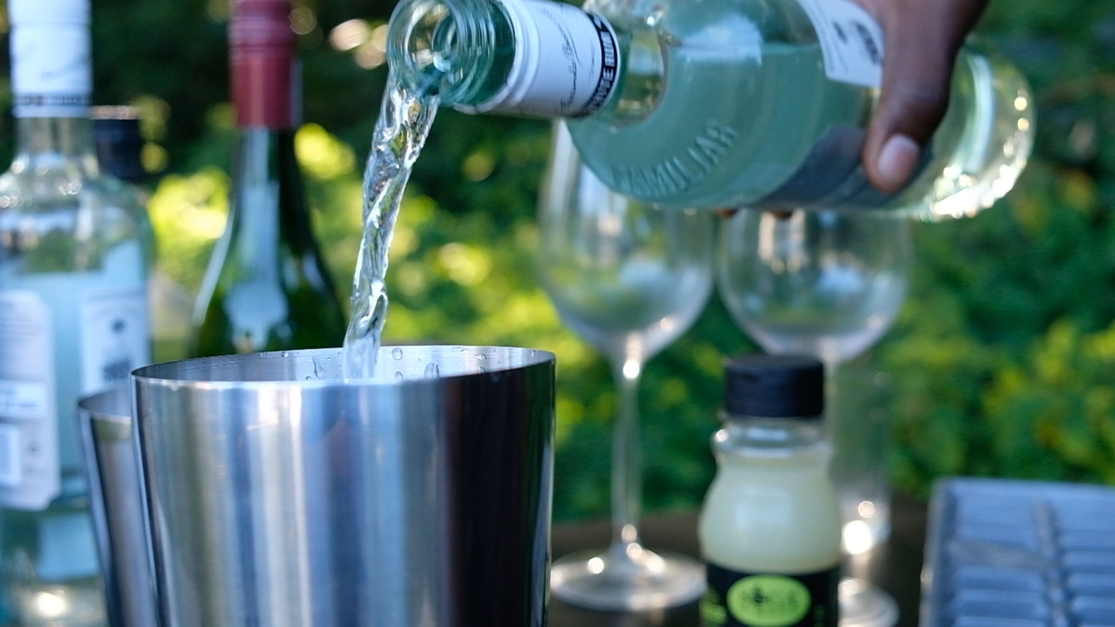 Clear spirit being poured into a metal cocktail shaker at an outdoor bar setup