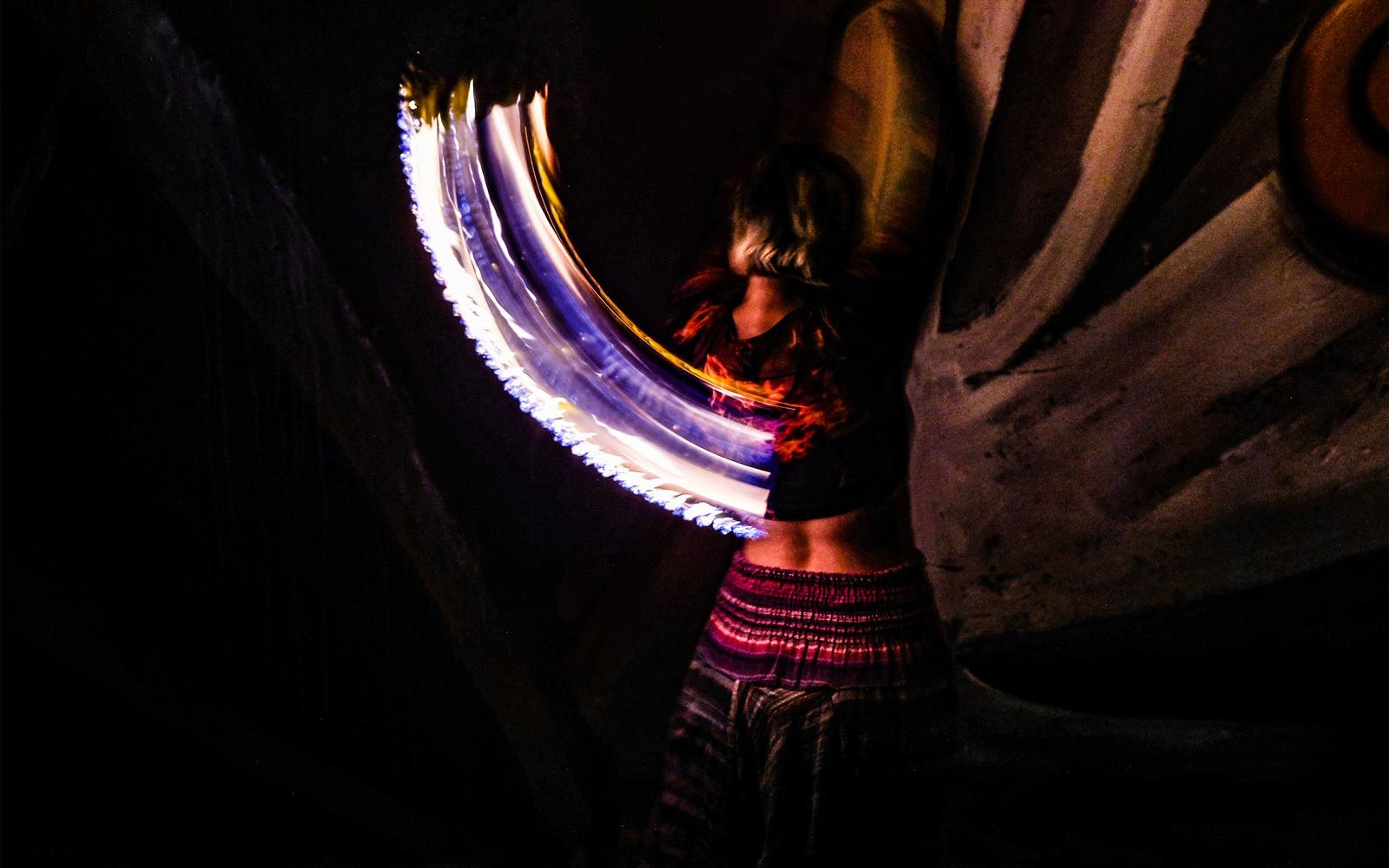 Motion-blurred performer with a purple and white light trail from a spinning fire staff, mural wall behind