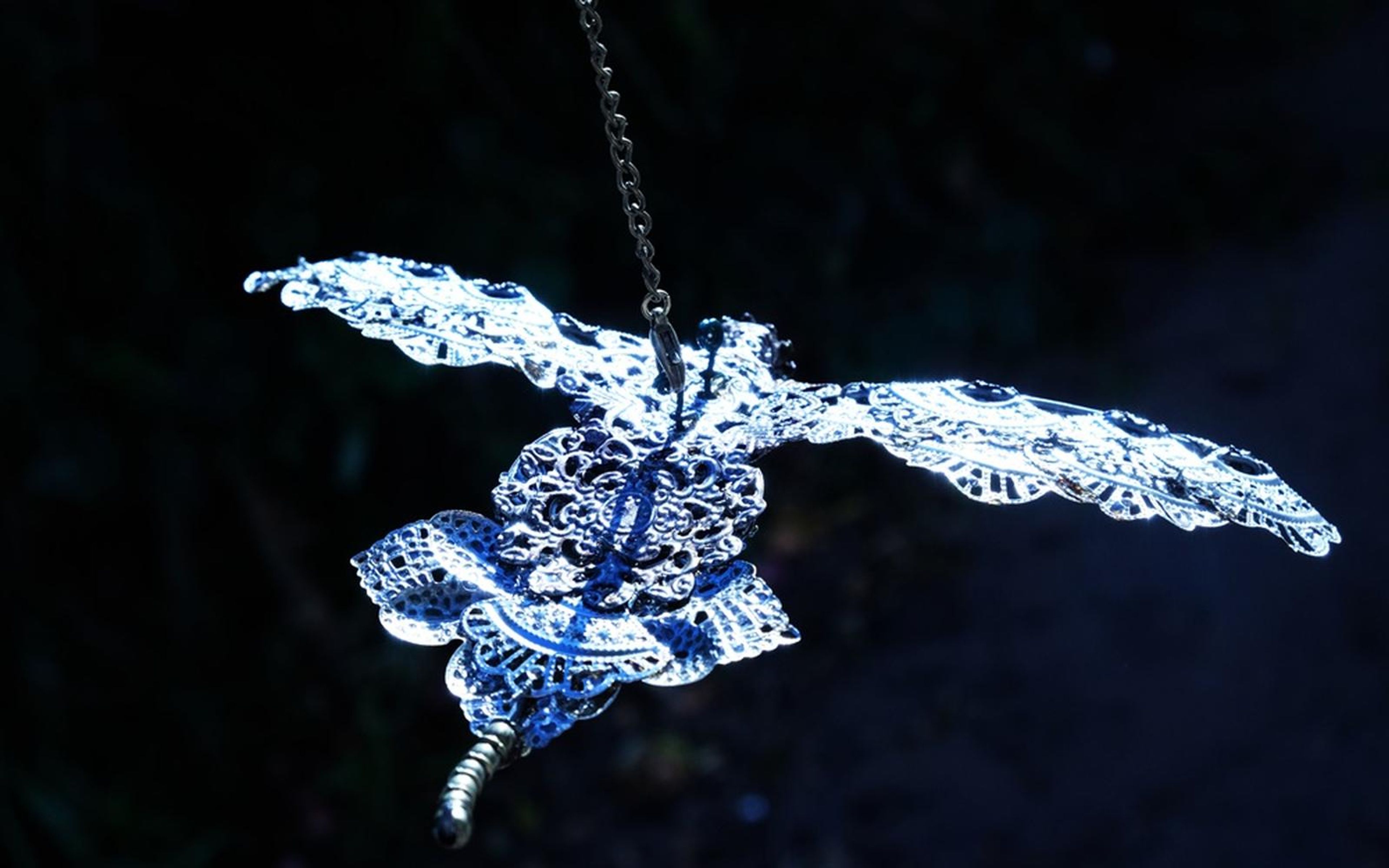 Glowing blue and white lace-like shape with outspread wings hanging from a chain in the dark