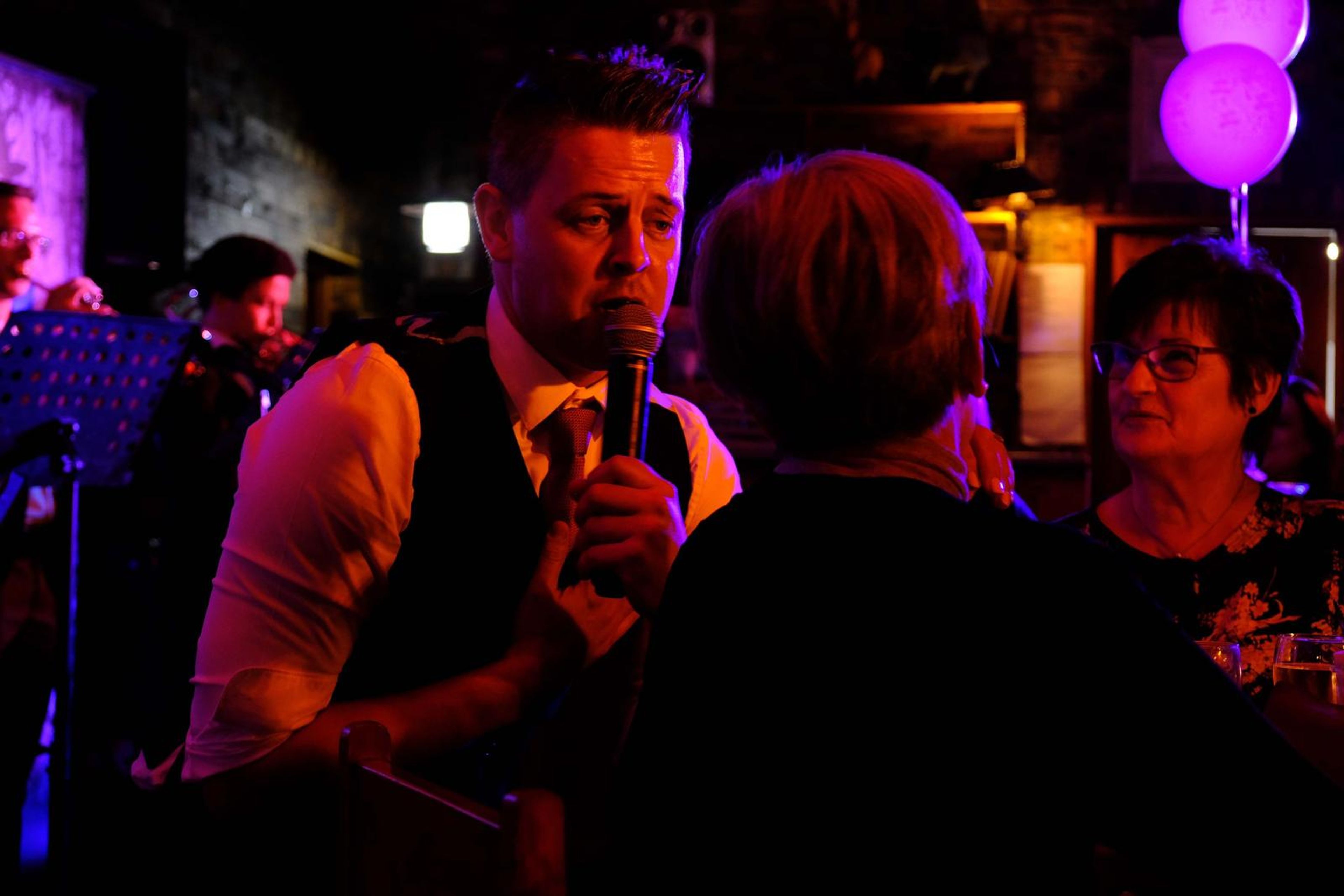 Singer in a waistcoat singing between tables of guests under balloons