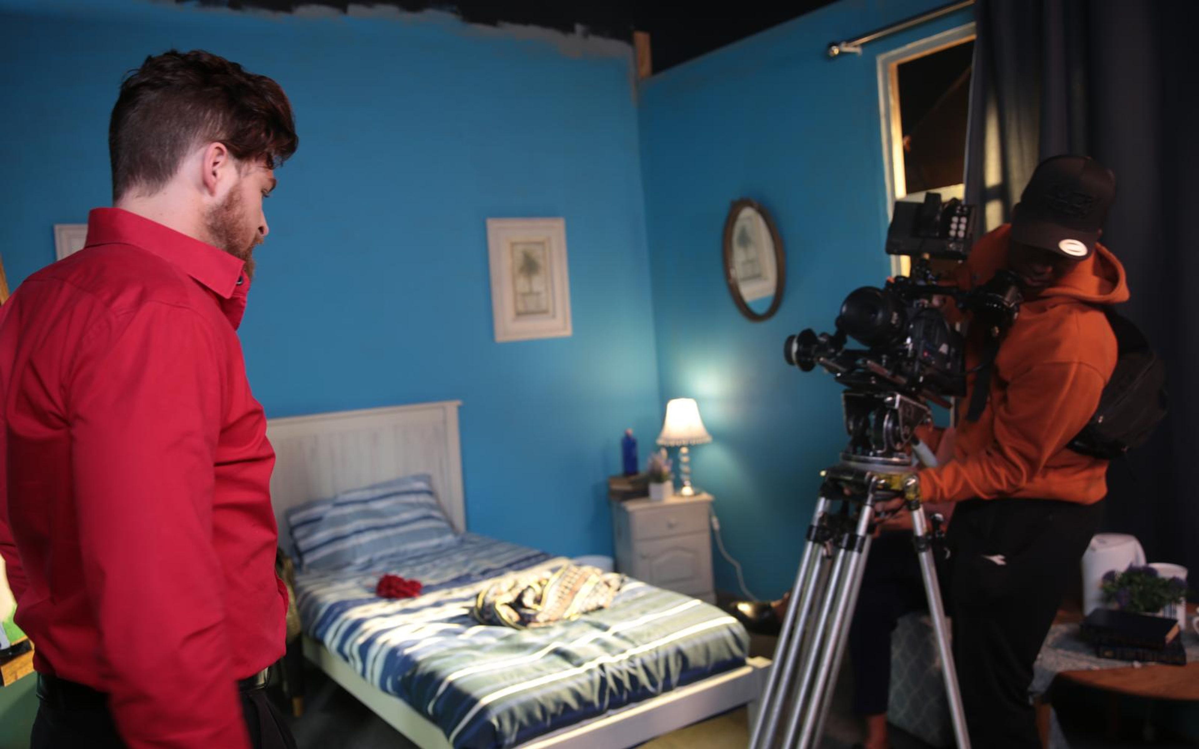 Man in a red shirt beside a bed on a blue bedroom set while a camera operator in an orange hoodie works behind a tripod