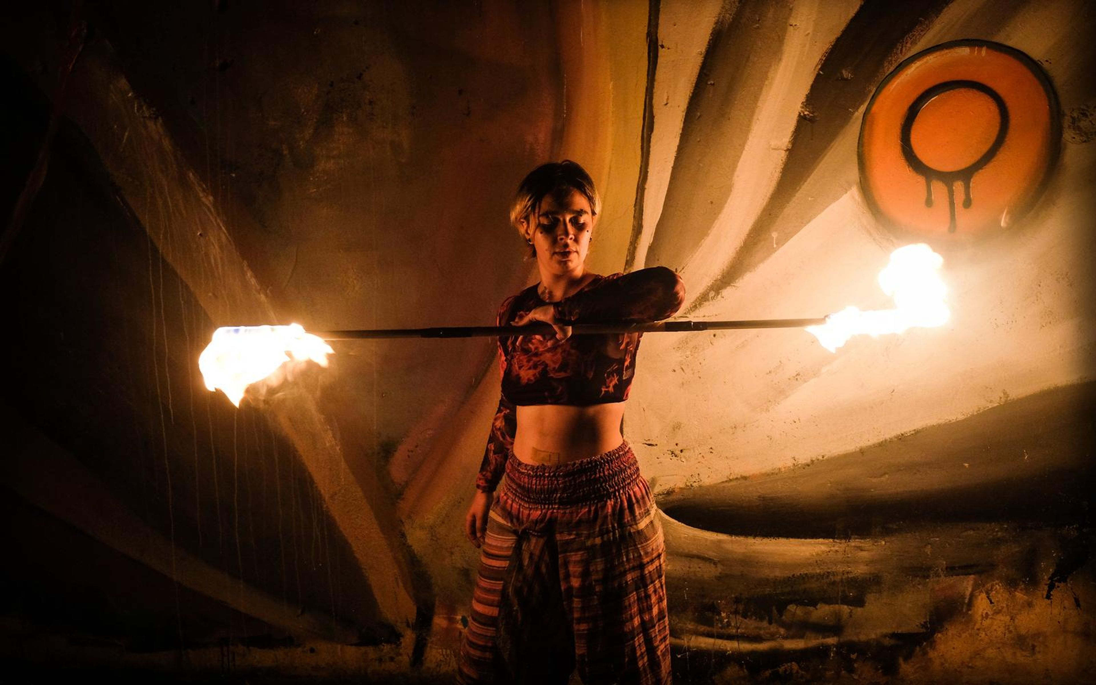 Performer in patterned trousers holding a fire staff horizontally in front of a mural with an orange circle emblem