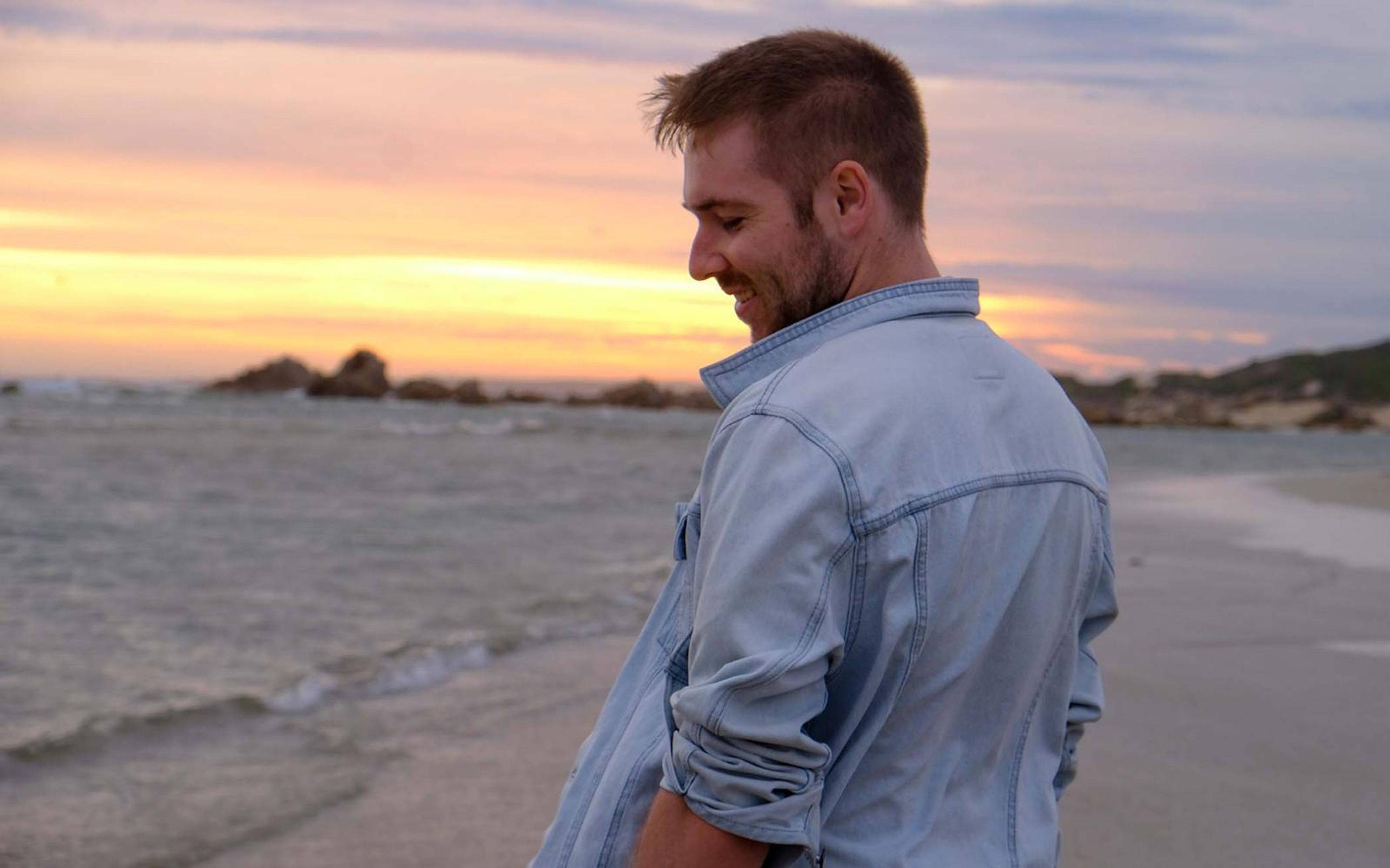 Man laughing with head bowed on a beach at dusk, soft pastel sky and distant rocks