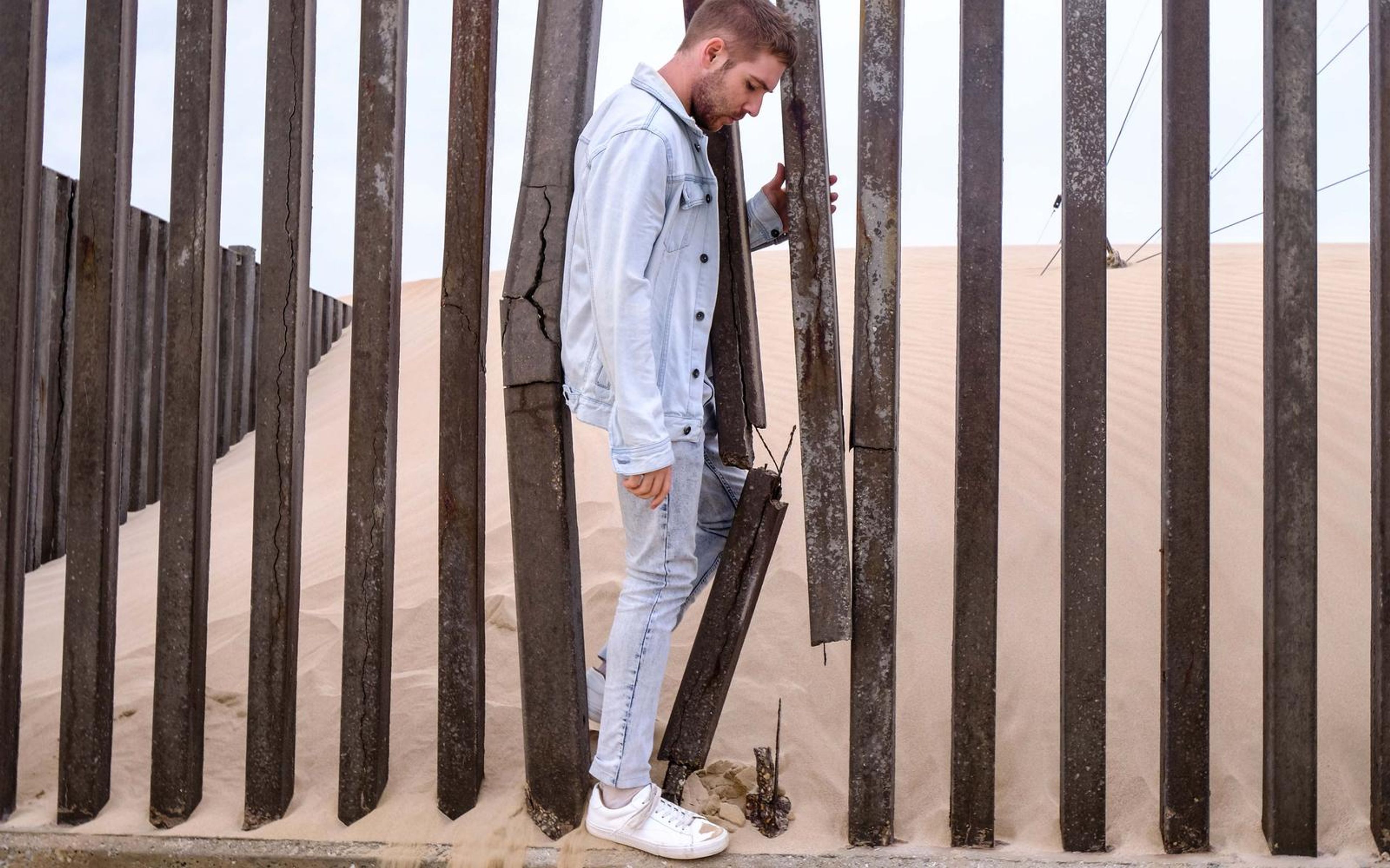Man in a light denim jacket leaning against tall wooden posts with sand behind them, white sneakers