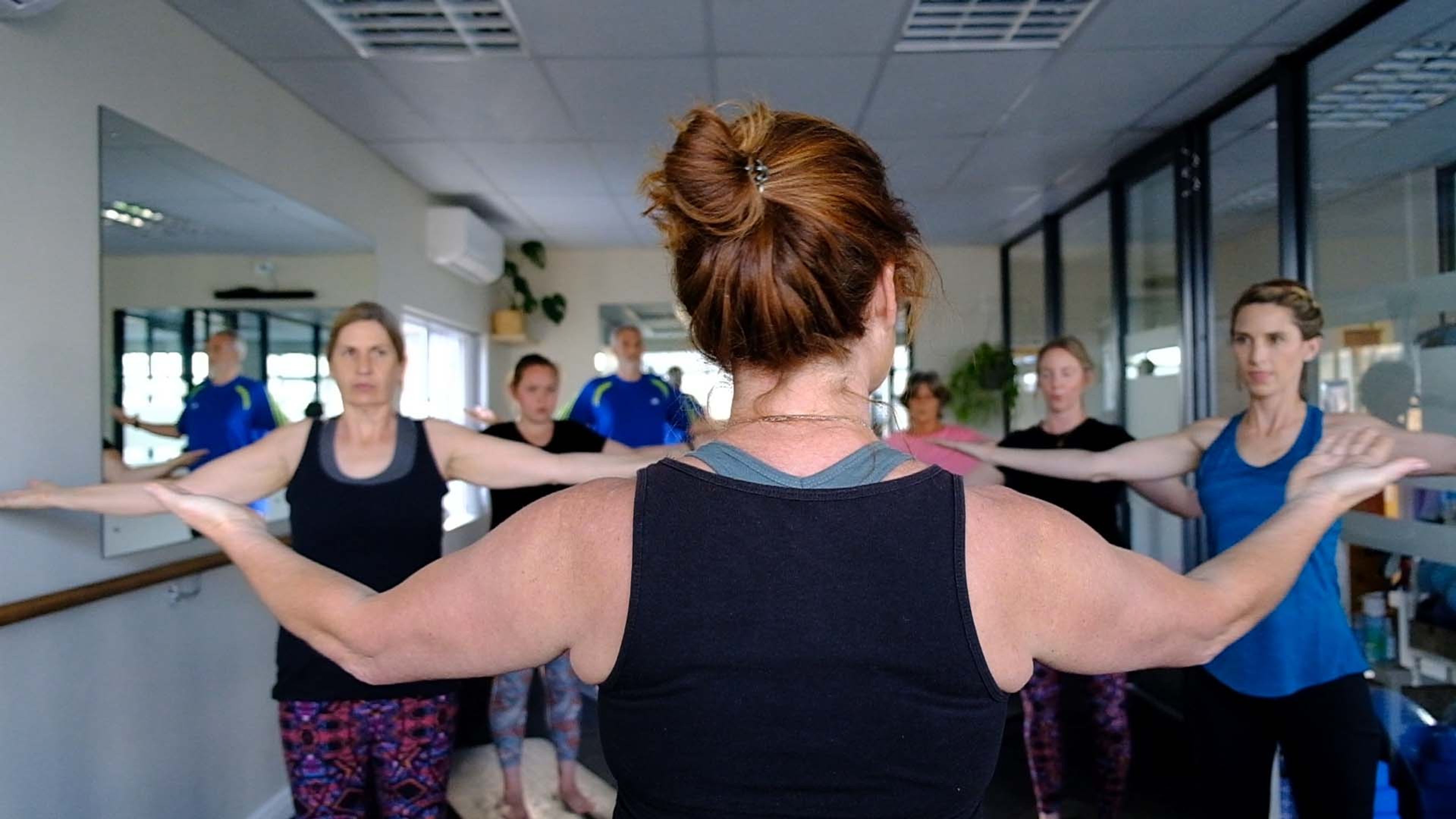 Rear view of an instructor with her arms held out wide, leading a group class standing in a row in front of a mirror