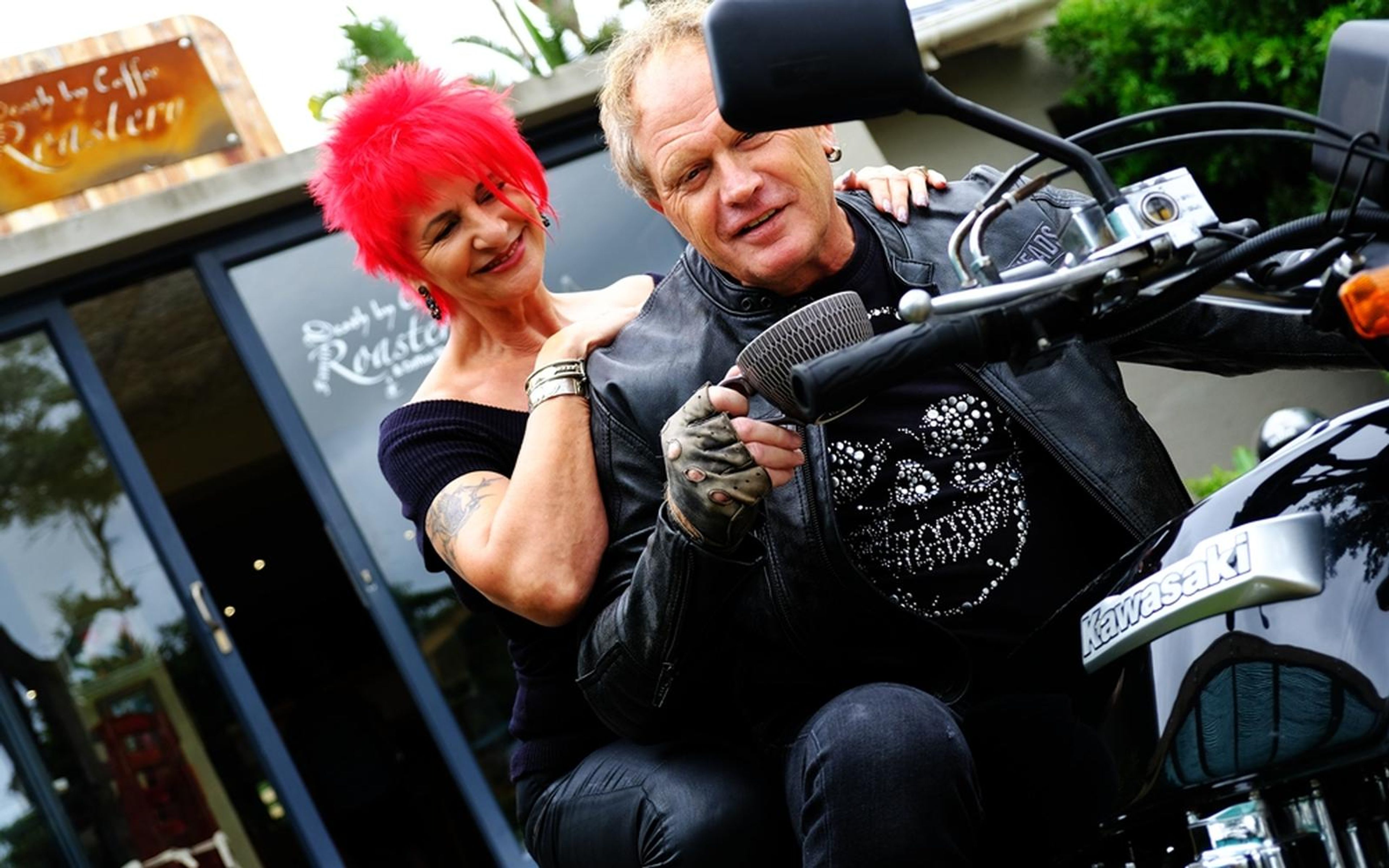 Woman with red hair resting her hand on a man's shoulder as they sit on a motorbike, both looking at a cup