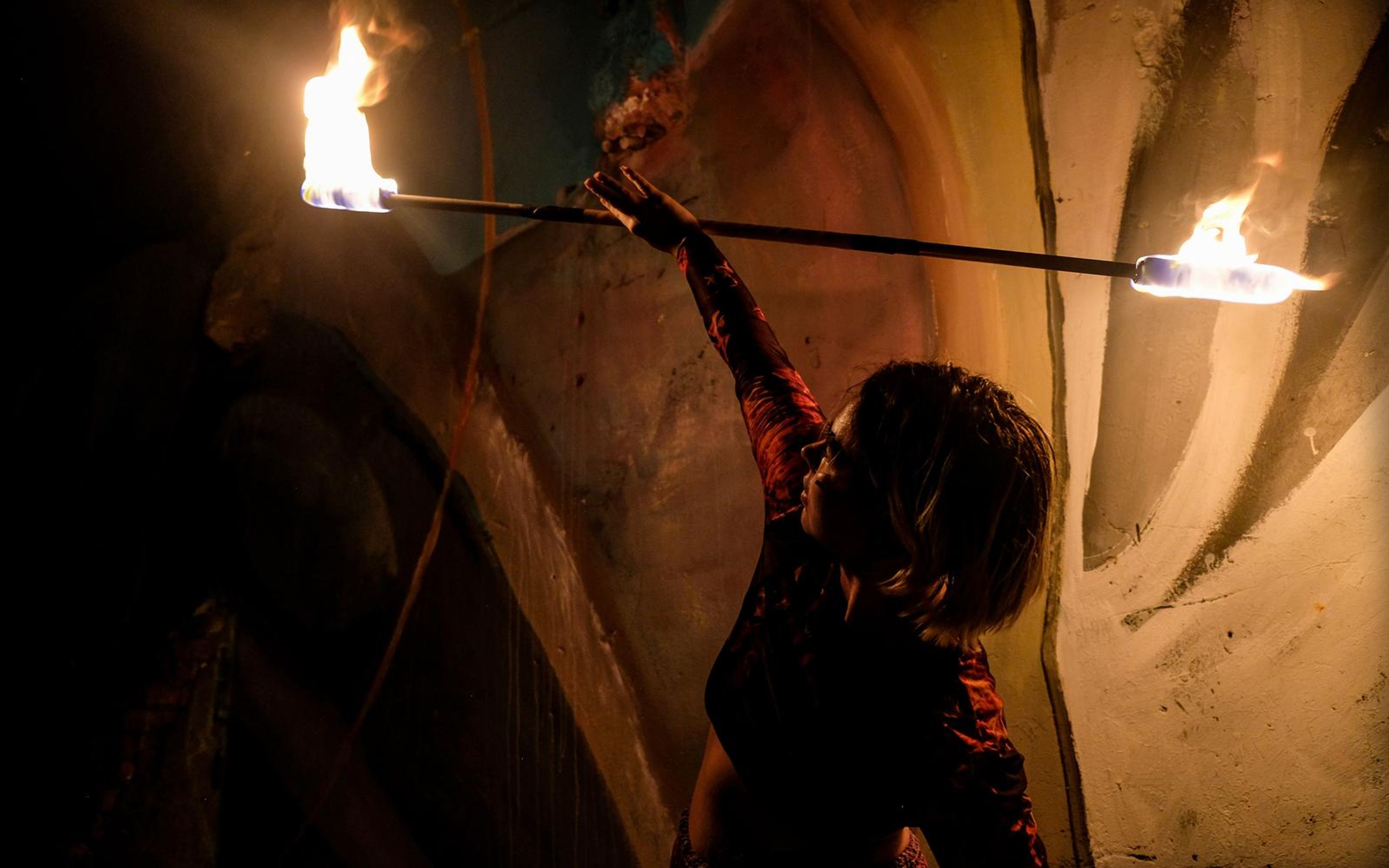Performer in profile at right holding a fire staff overhead with flames at both ends against a warm wall