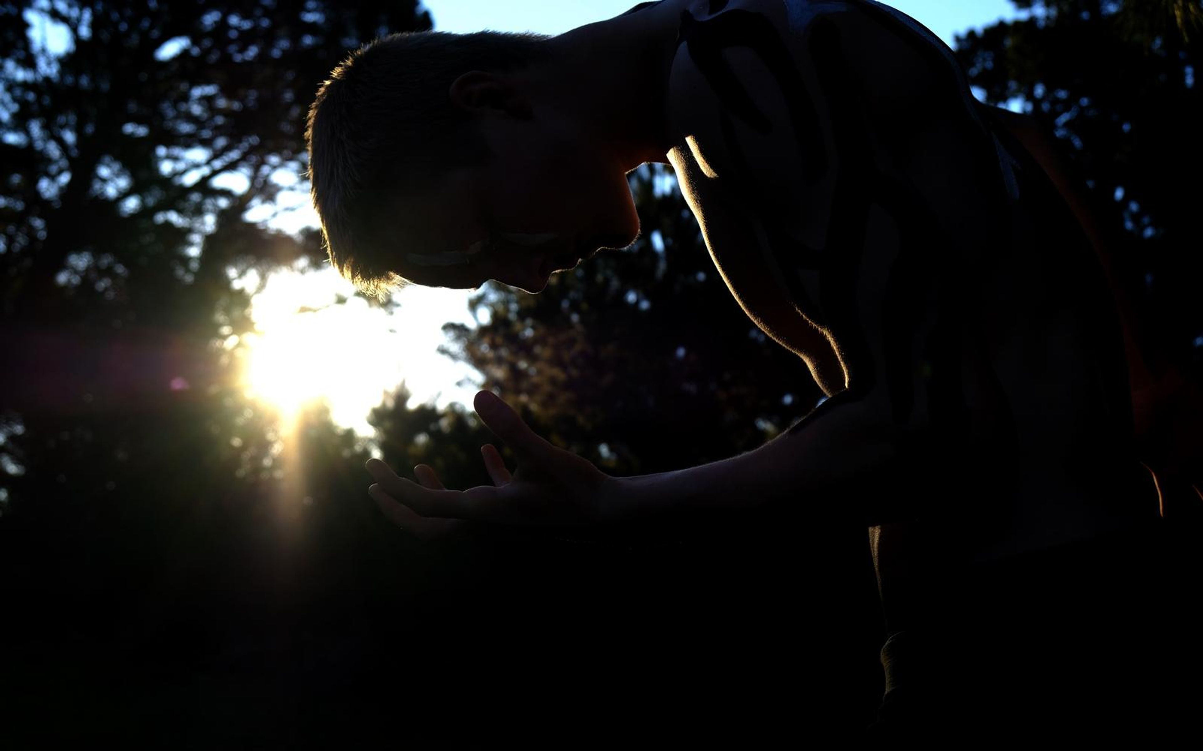Performer bowed forward in silhouette with sun flare between the trees and a hand reaching out
