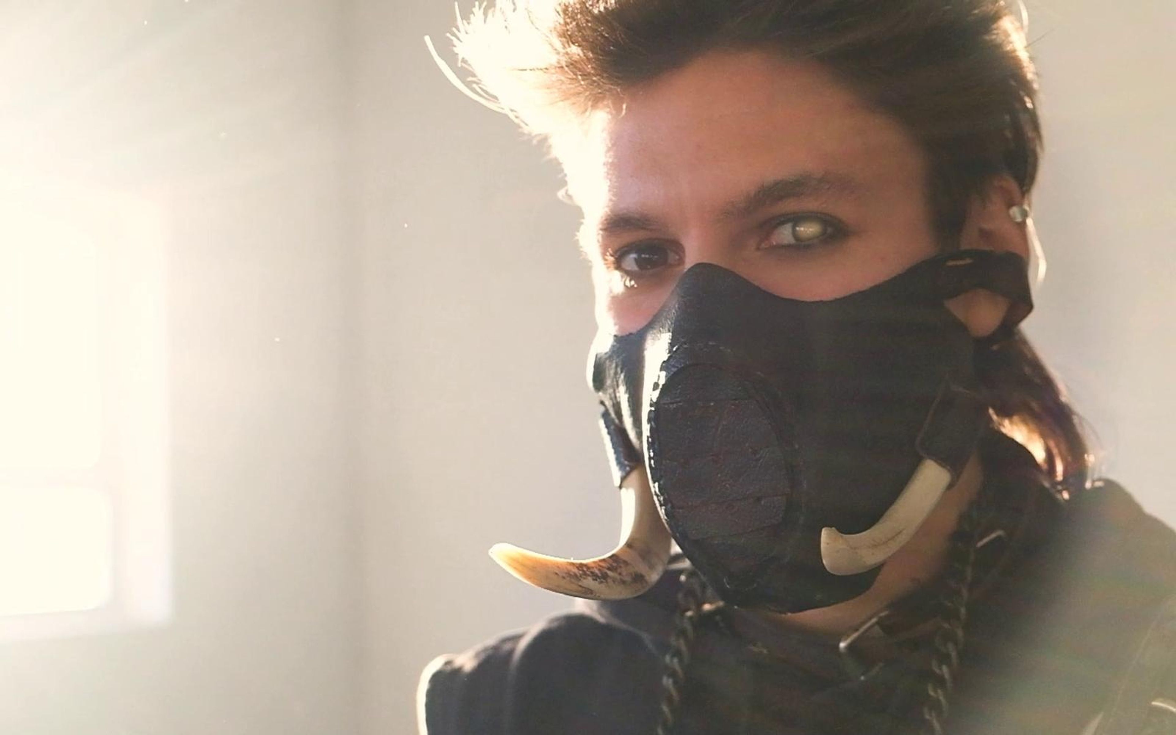 Close view of the masked warrior's eyes above a studded leather mask with curved tusks, glowing in warm flare light
