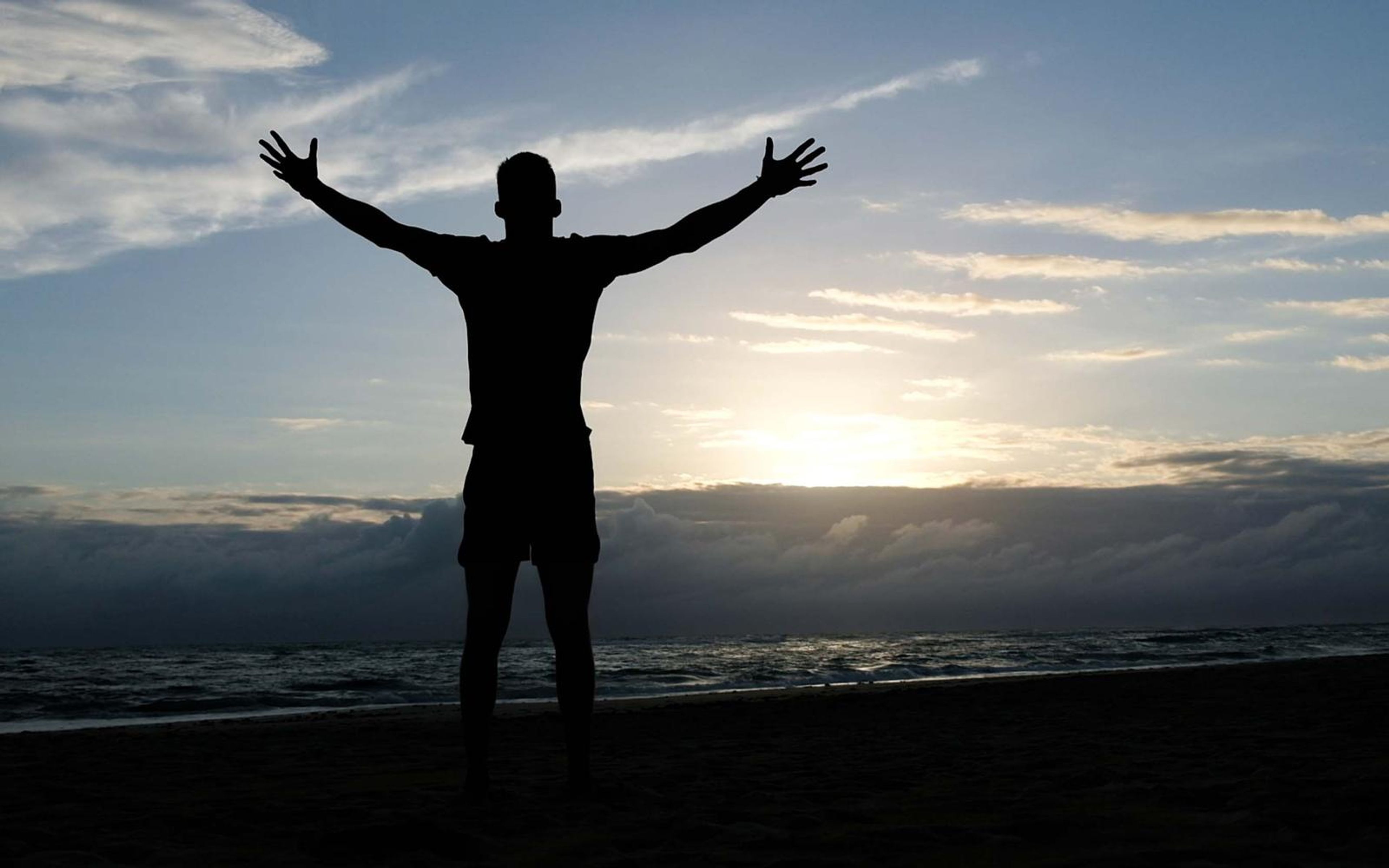 Silhouetted man standing with arms spread wide on a beach at dusk under layered clouds