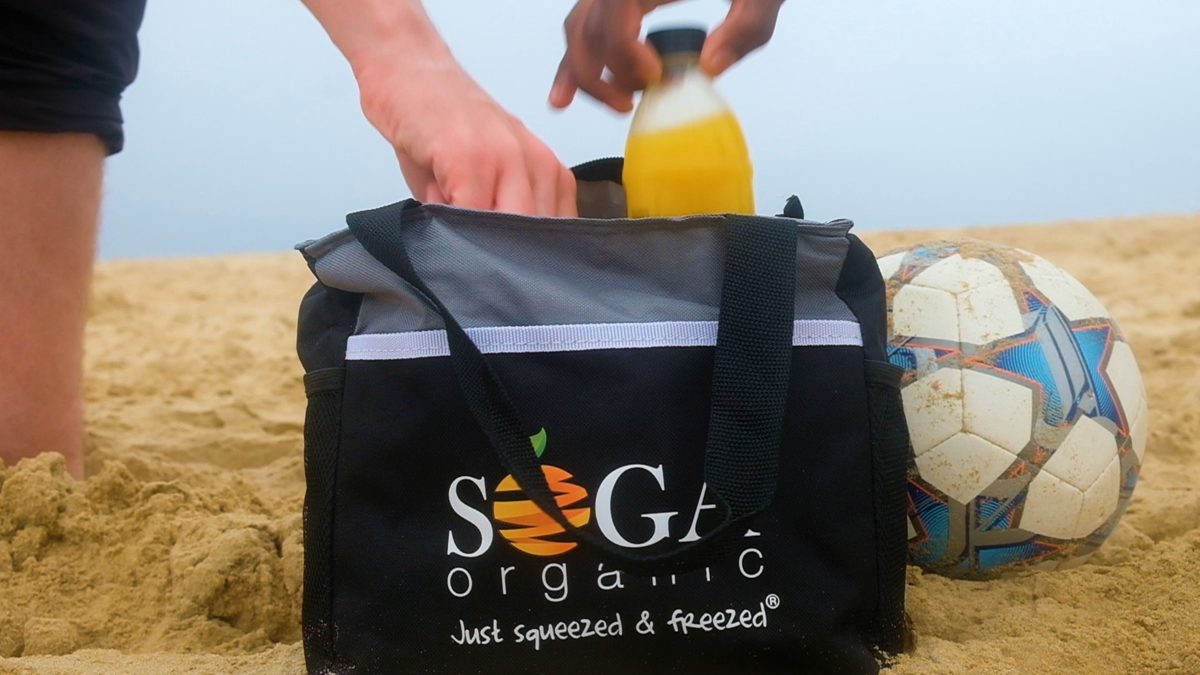 SOGA Organic cooler bag on the beach with a bottle of orange juice and a football