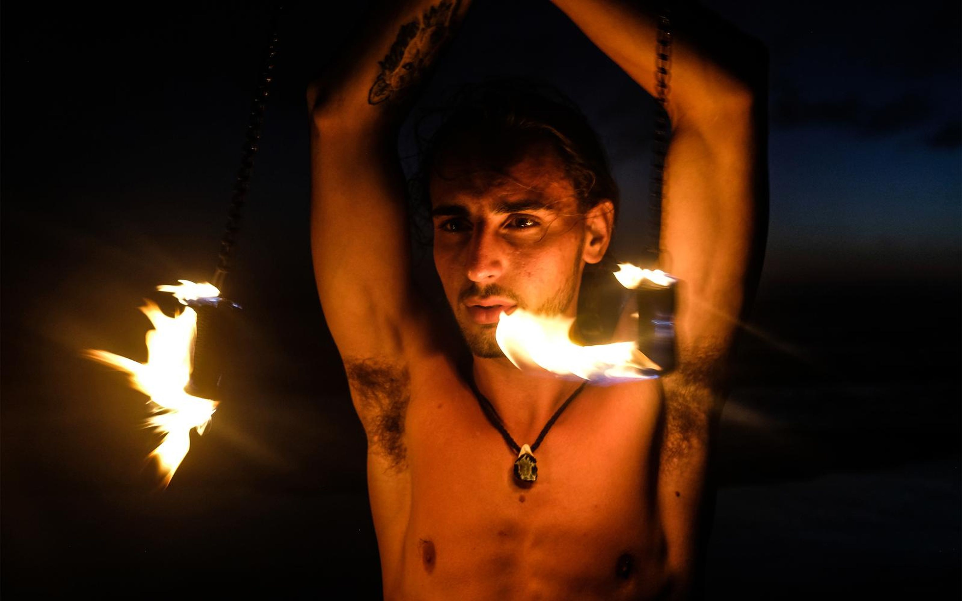 Man looking toward the camera with fire poi burning near his chin and shoulder, dusk sky