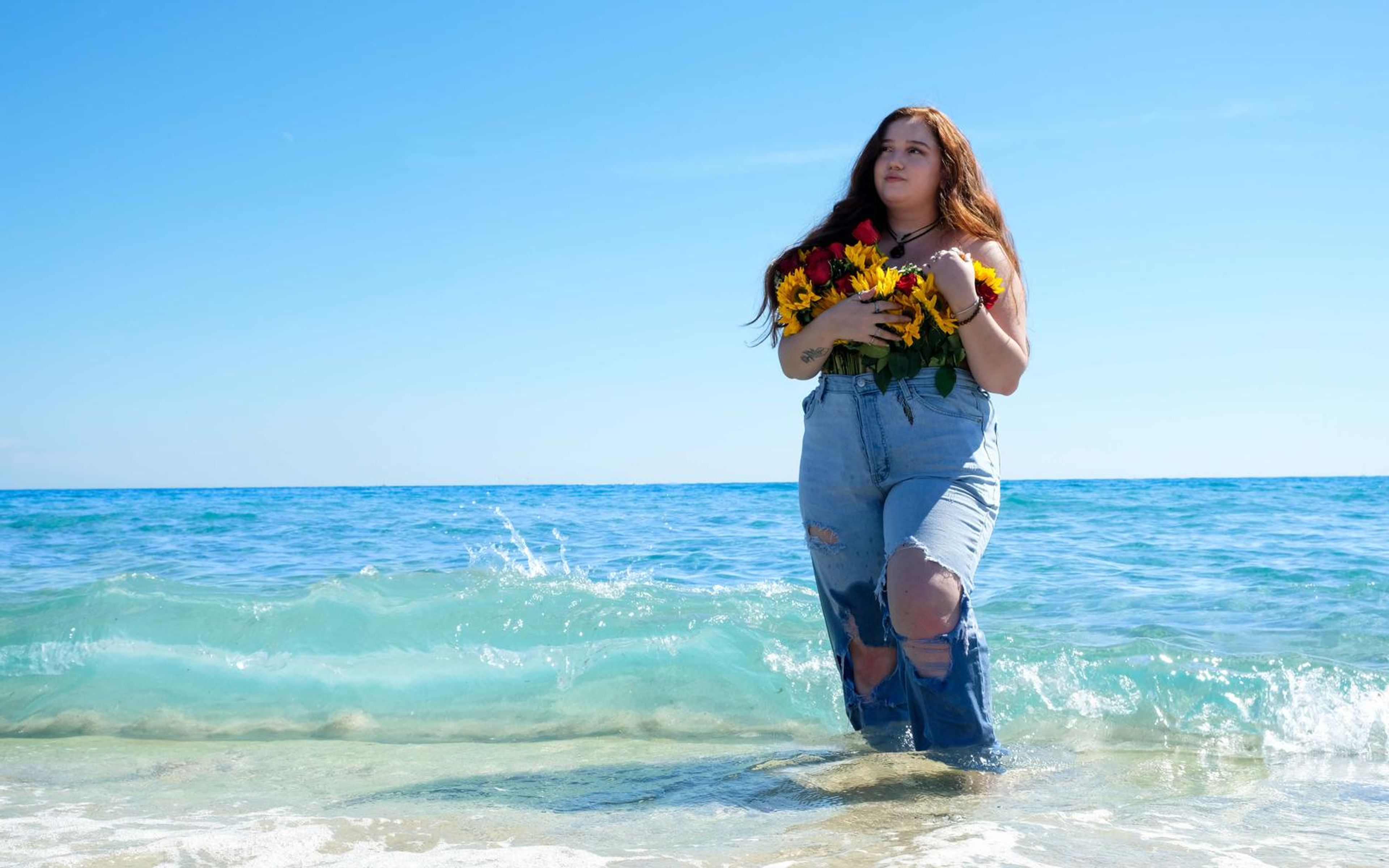 Model standing in clear shallow water near the shore, bouquet to chest, soft turquoise wave behind
