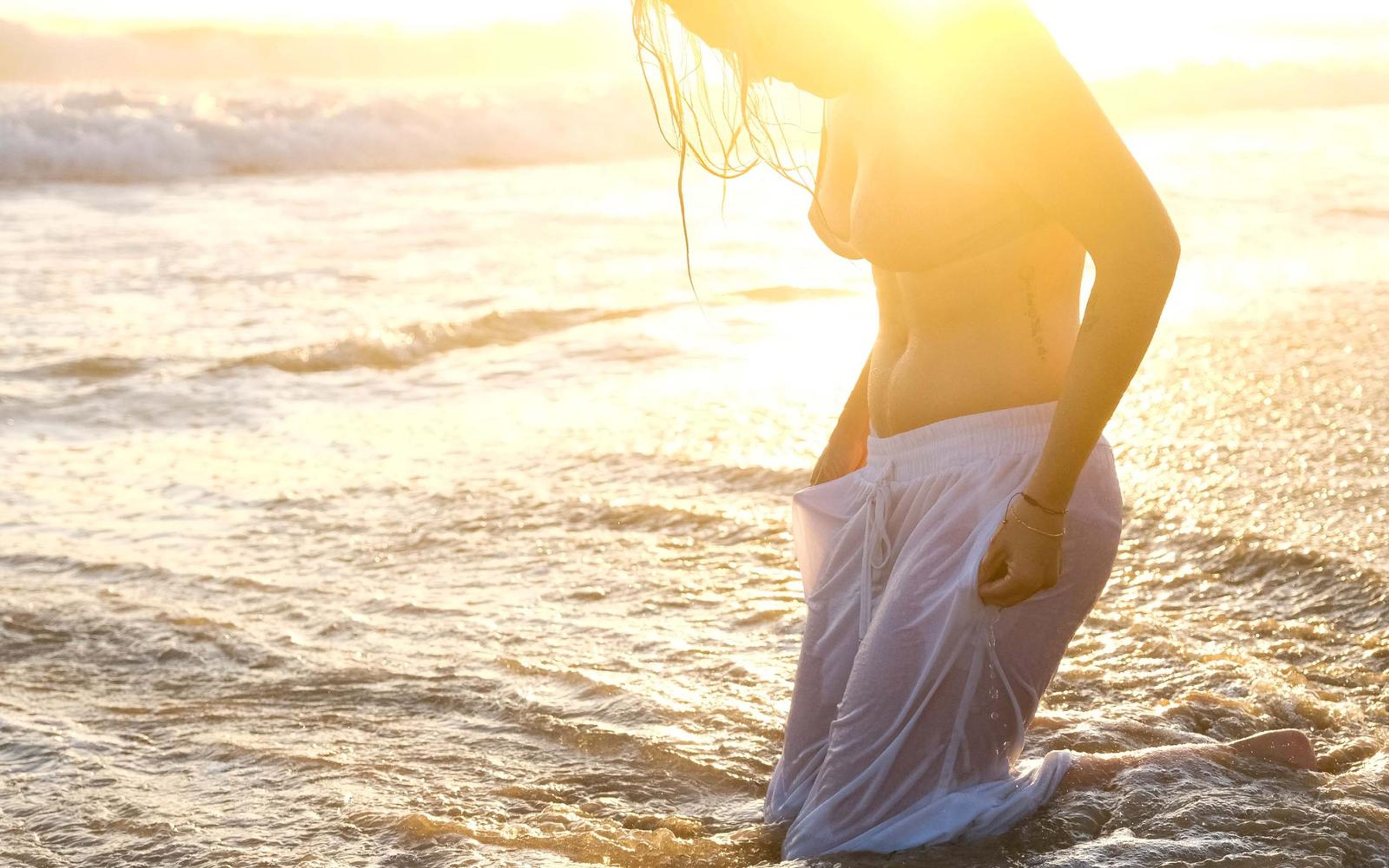Woman standing in knee-deep surf with head bowed in strong sunlight, wet hair falling, white trousers clinging