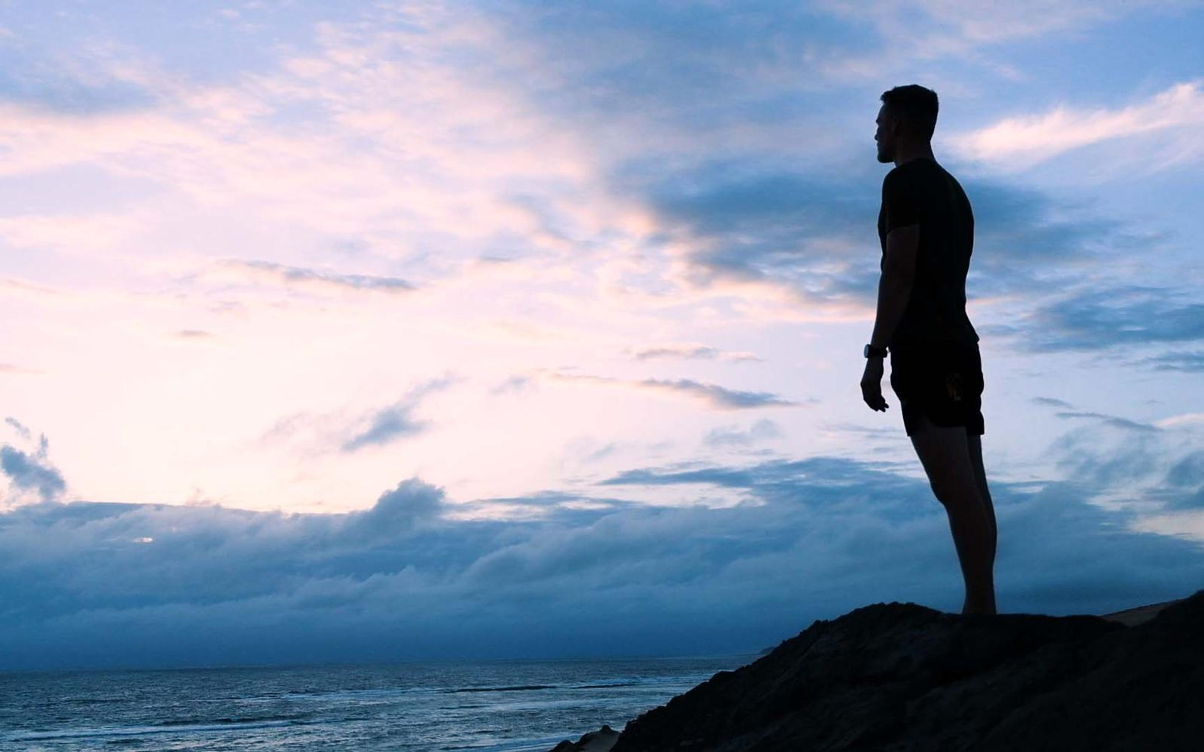 Silhouette of a man standing on a rocky outcrop above the sea under a pink and blue dusk sky