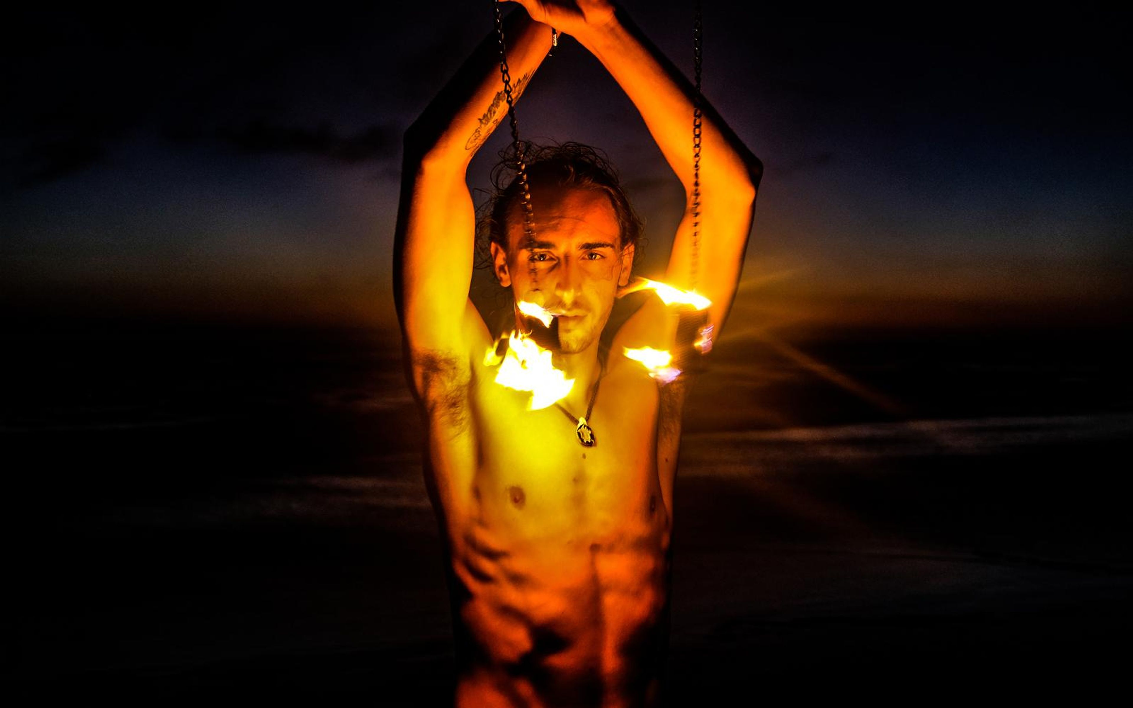 Man with a golden-lit face between raised arms and flames at both sides, sunset sky and sea behind