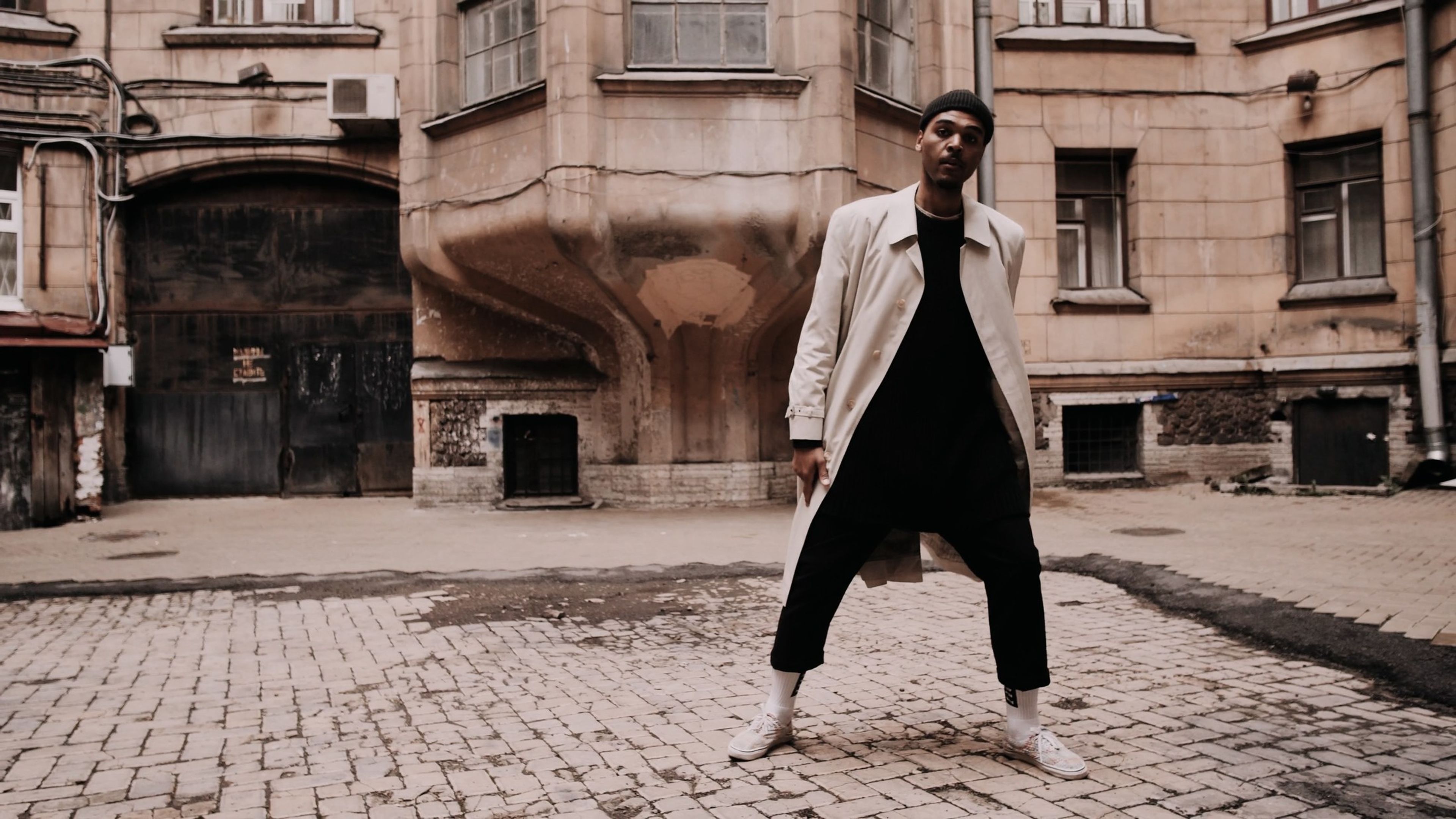 Man in a long cream coat and white sneakers standing in a brick courtyard