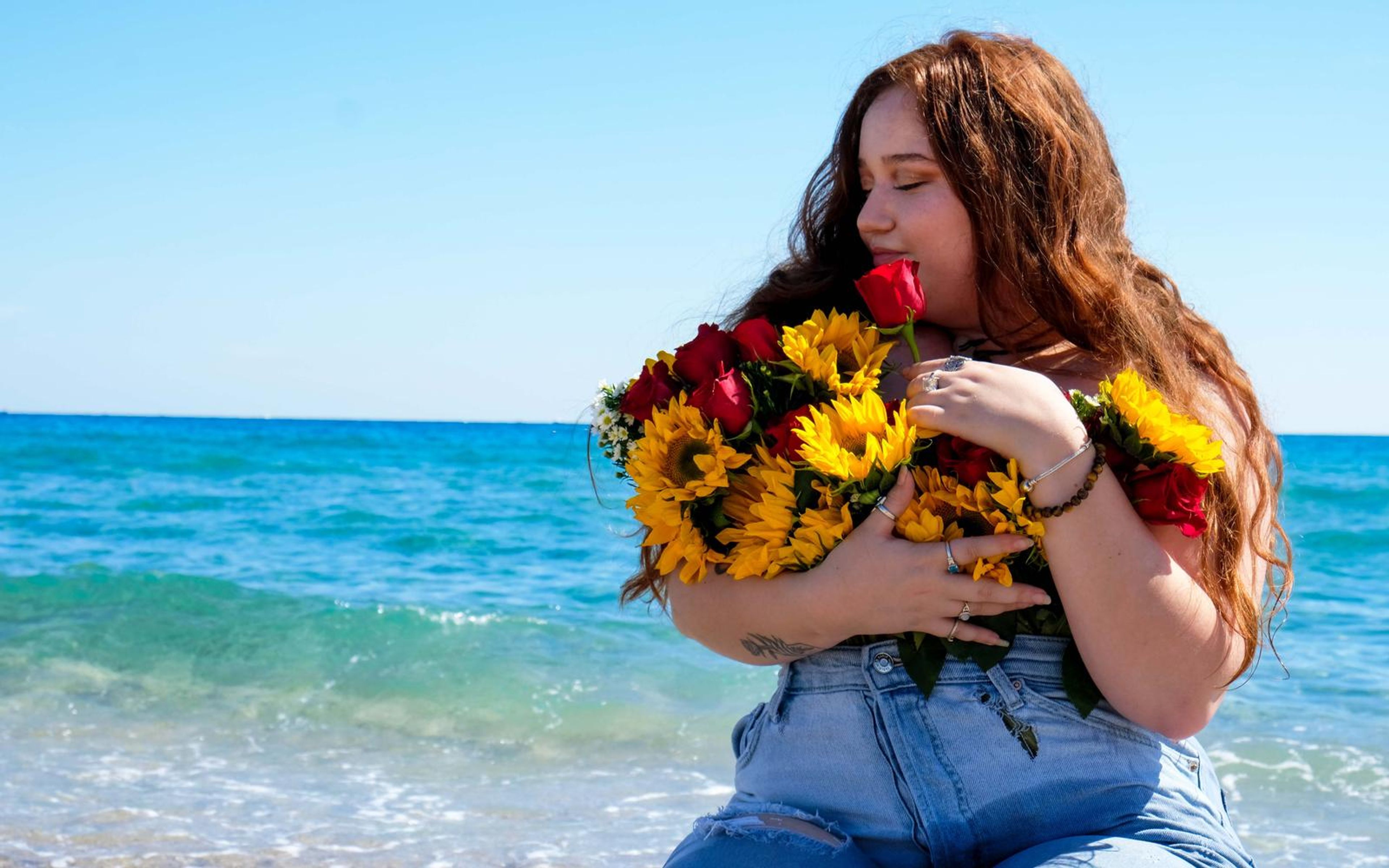 Woman seated by the shoreline smiling down at a red rose, arms wrapped around a bouquet of sunflowers, roses and daisies, turquoise waves behind