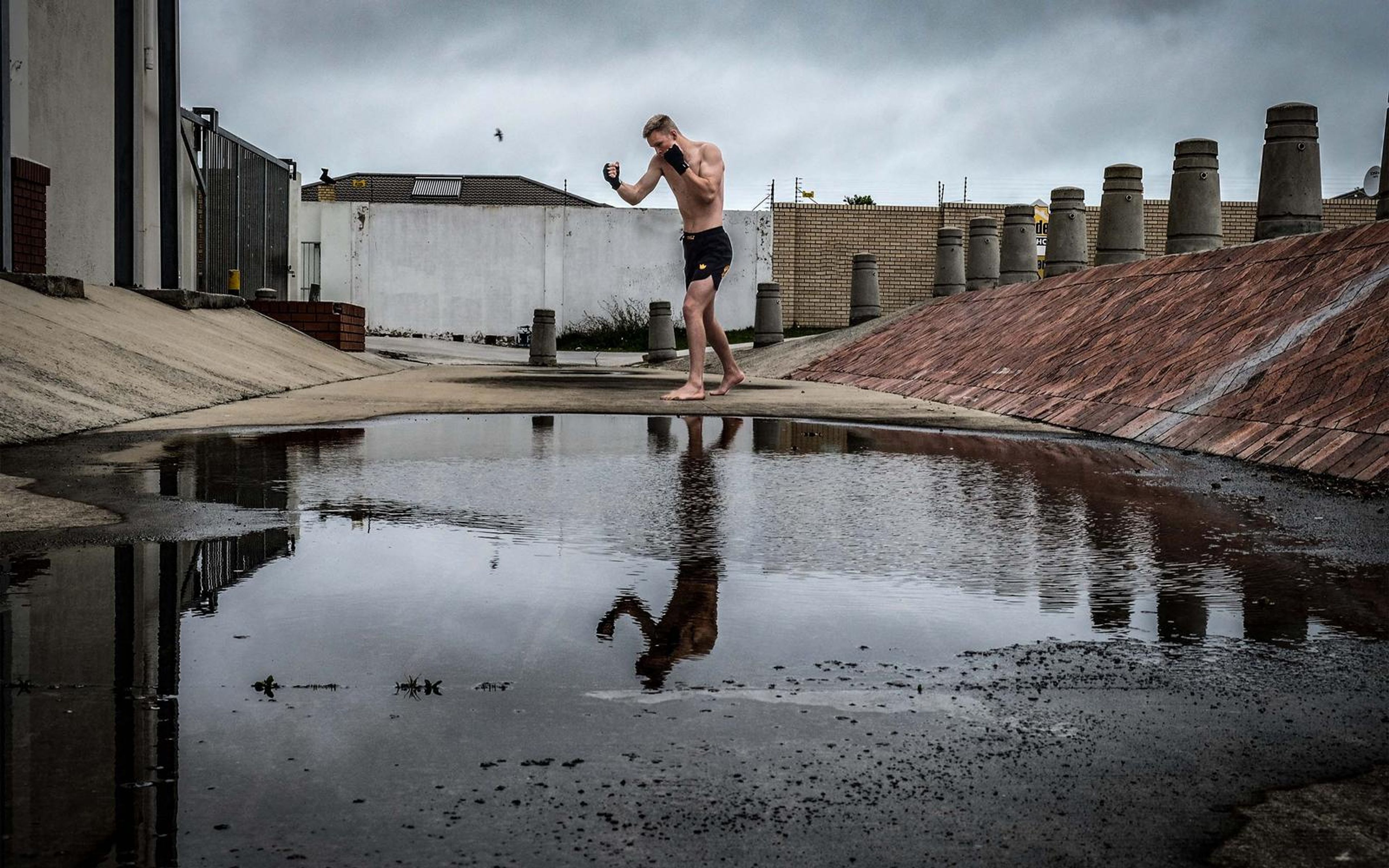 Man in a guard pose with one hand raised, reflected in the puddle under dark storm clouds