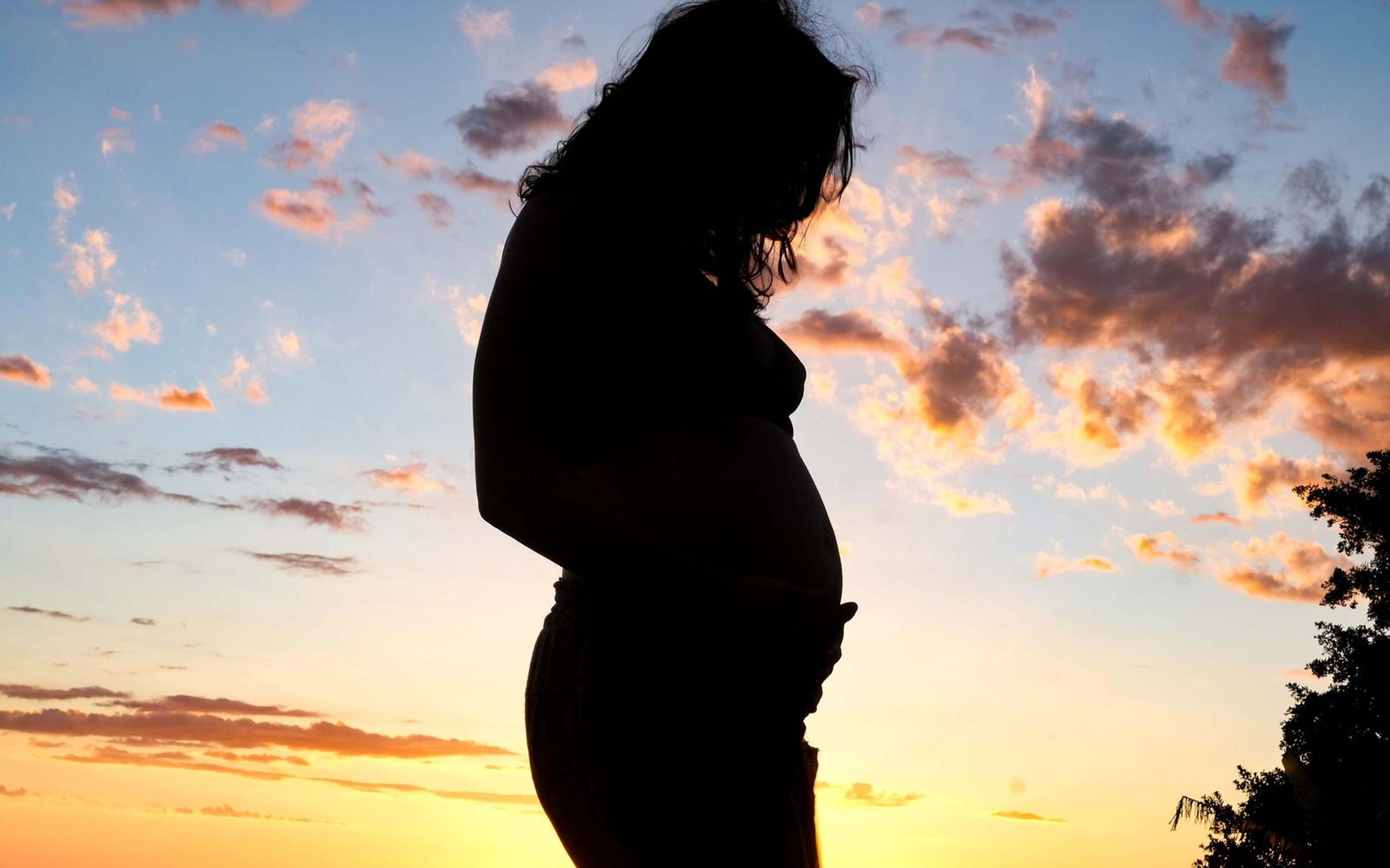 Pregnant woman in silhouette with head bowed, hand at her belly, colourful dusk clouds