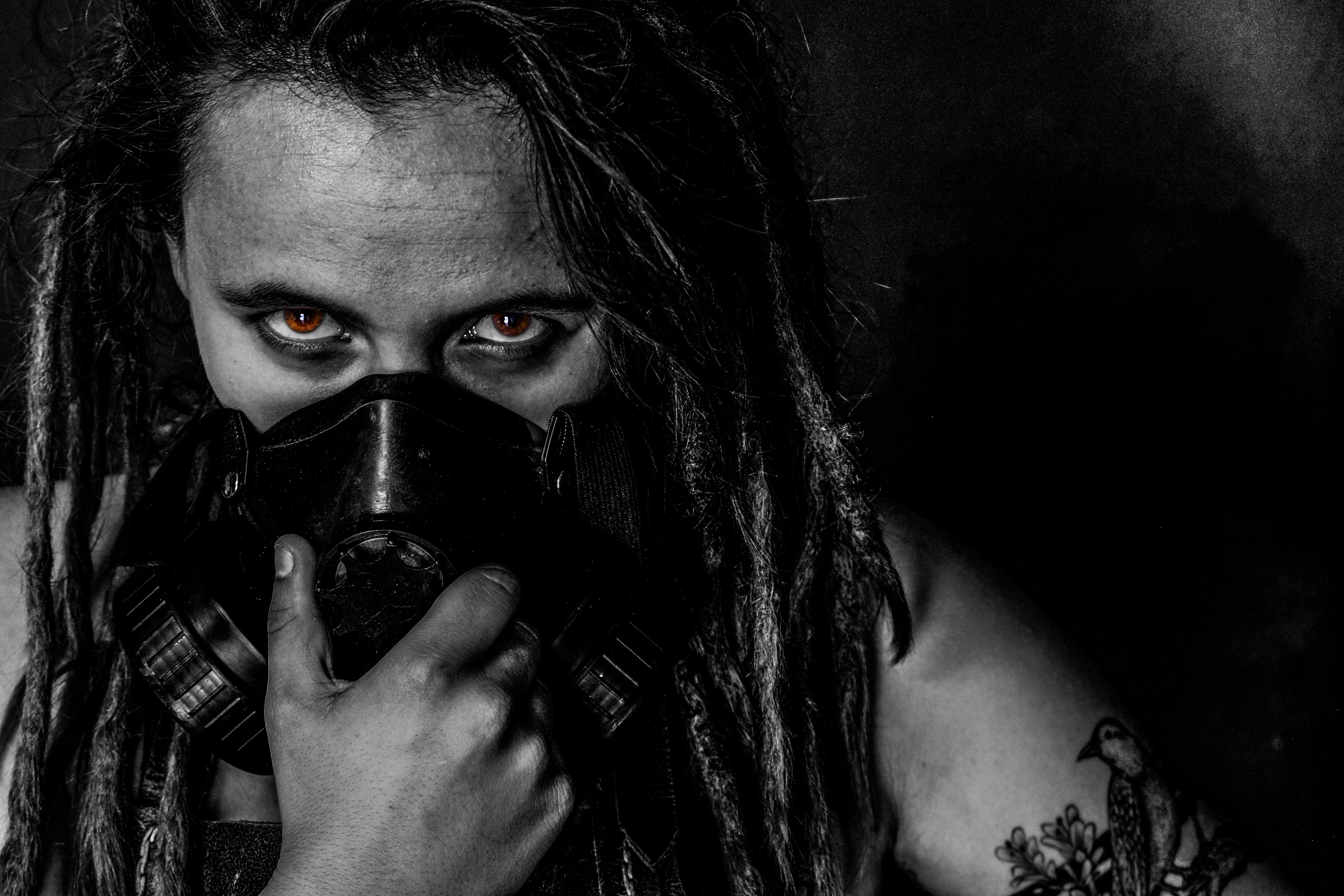 Black and white portrait, subject wearing a gas mask with red-tinted eyes, dreadlocks falling across the face