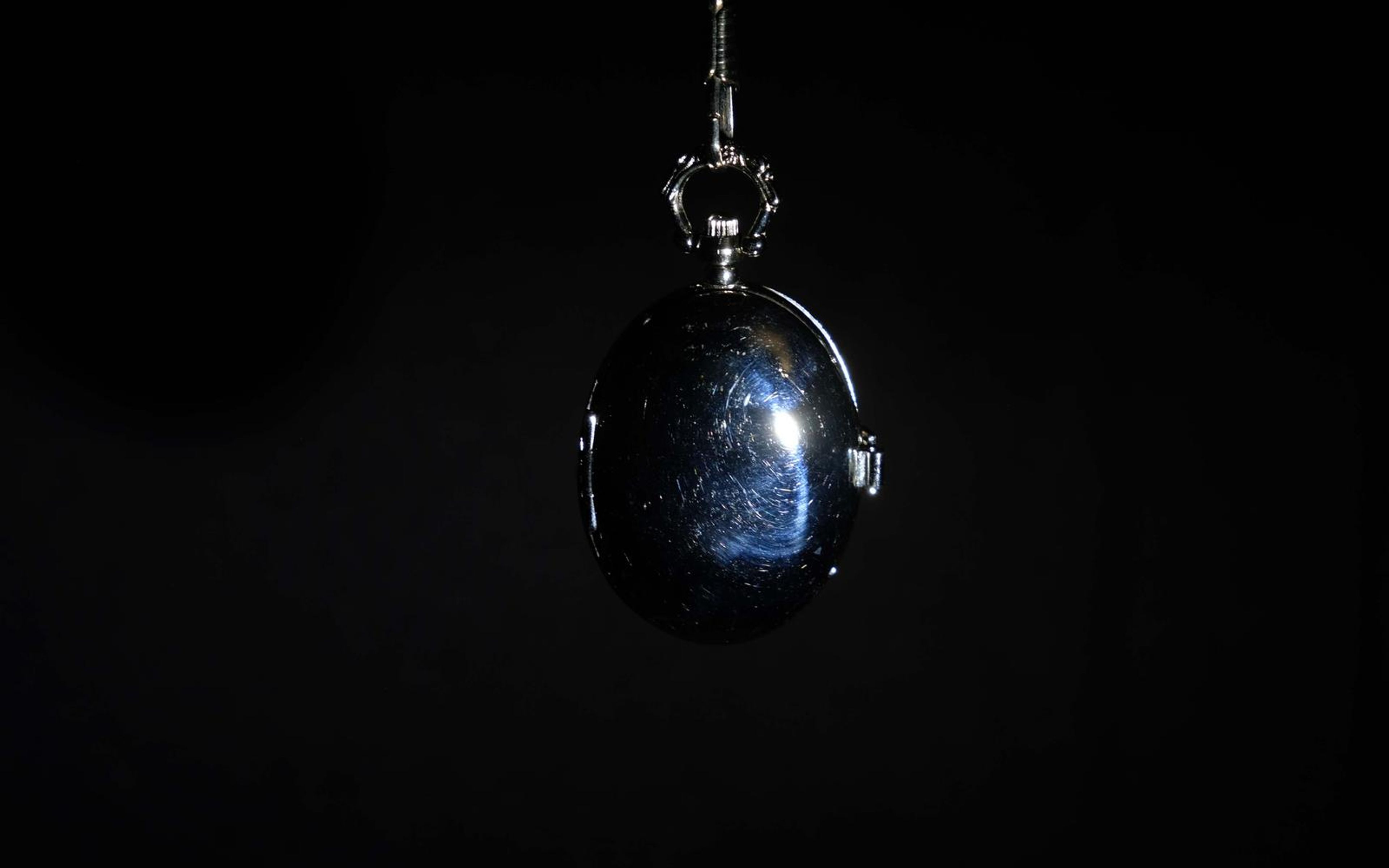 Silver pocket watch case hanging from a chain against black, reflecting a blue-white highlight