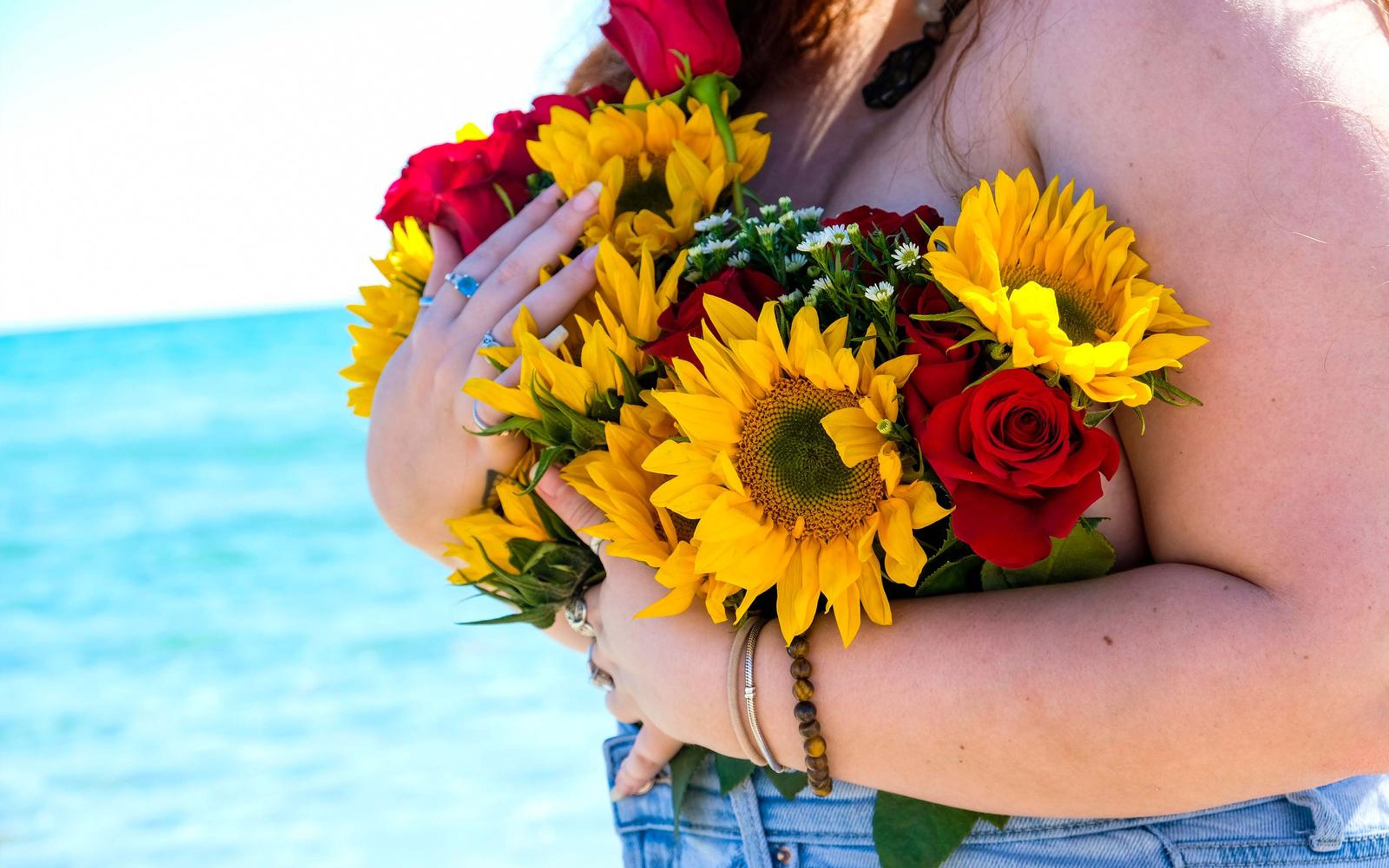 Close view of arms cradling a sunflower and rose bouquet, rings and bracelets visible, denim waistband at the bottom, bright cyan sea behind