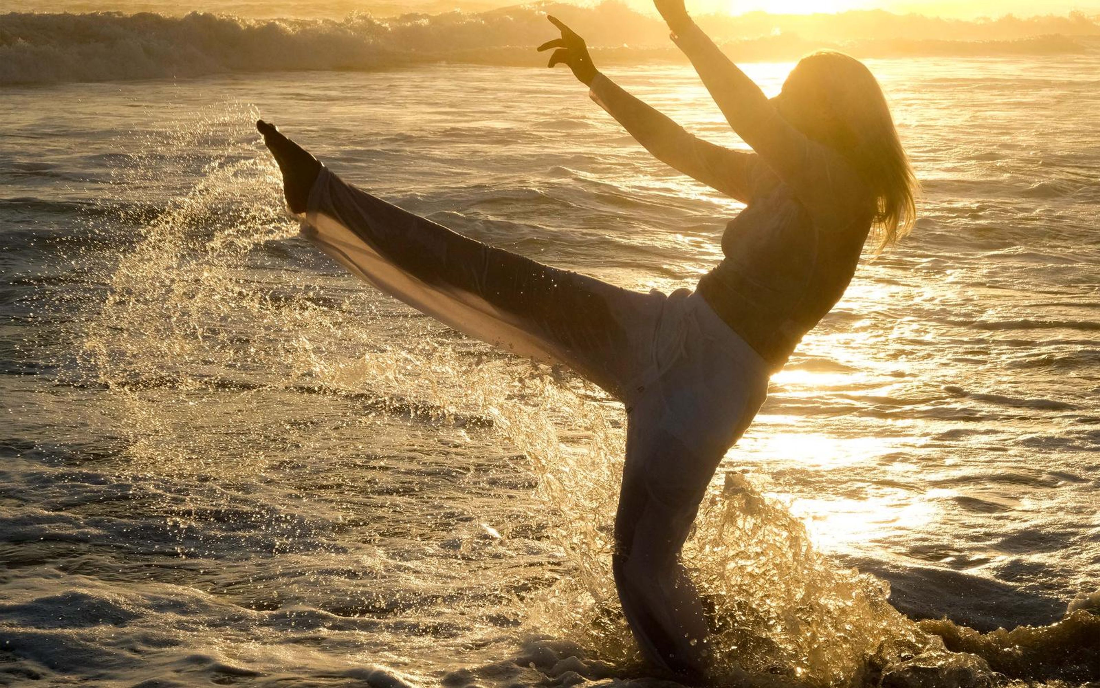 Woman kicking a leg high at the water's edge, droplets lit gold by the setting sun