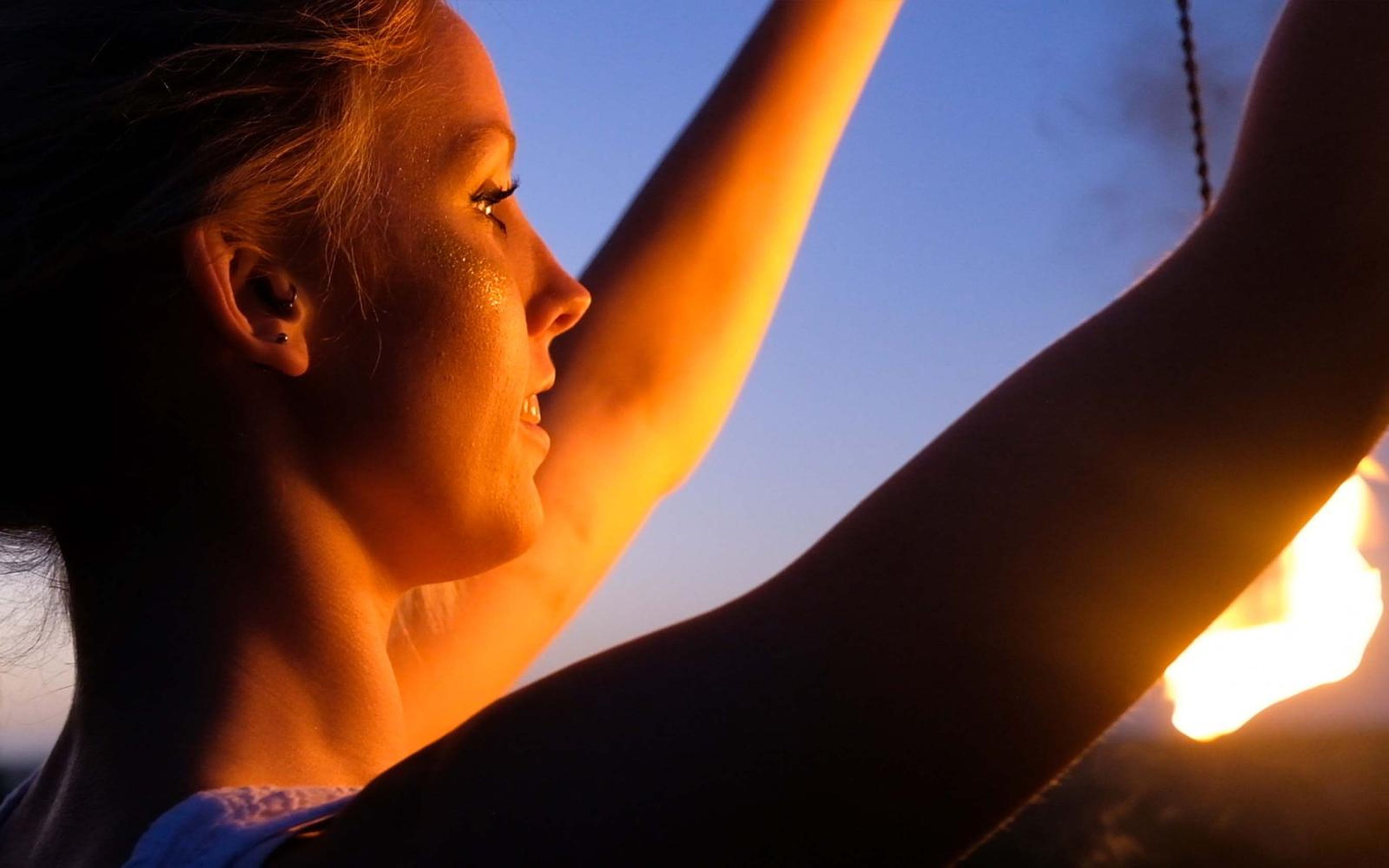 Woman in profile with arms raised and a flame glowing at the lower right against a blue dusk sky