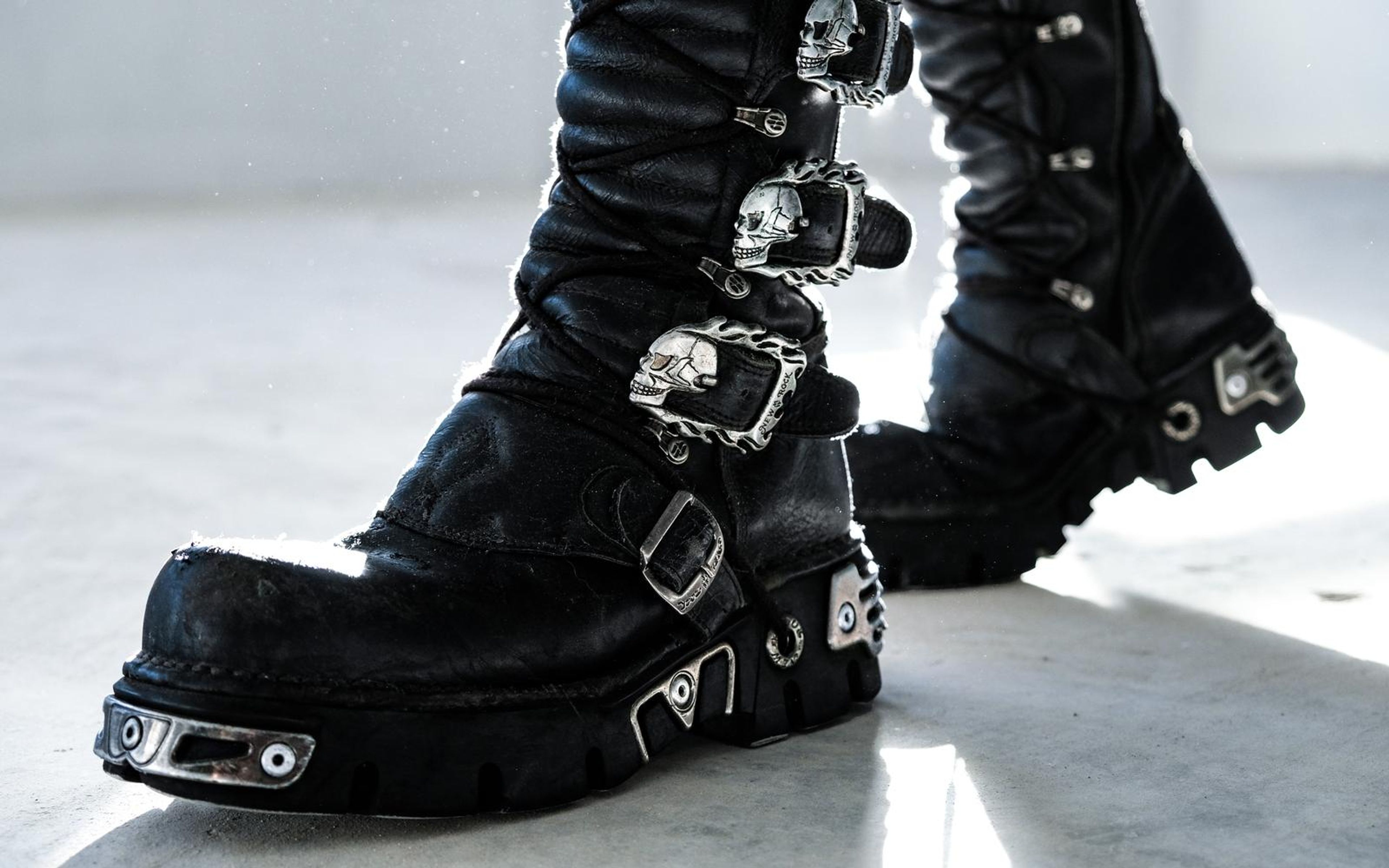 Black boot with skull-shaped clasps lifted mid-step, strong backlight and shadows on a concrete floor