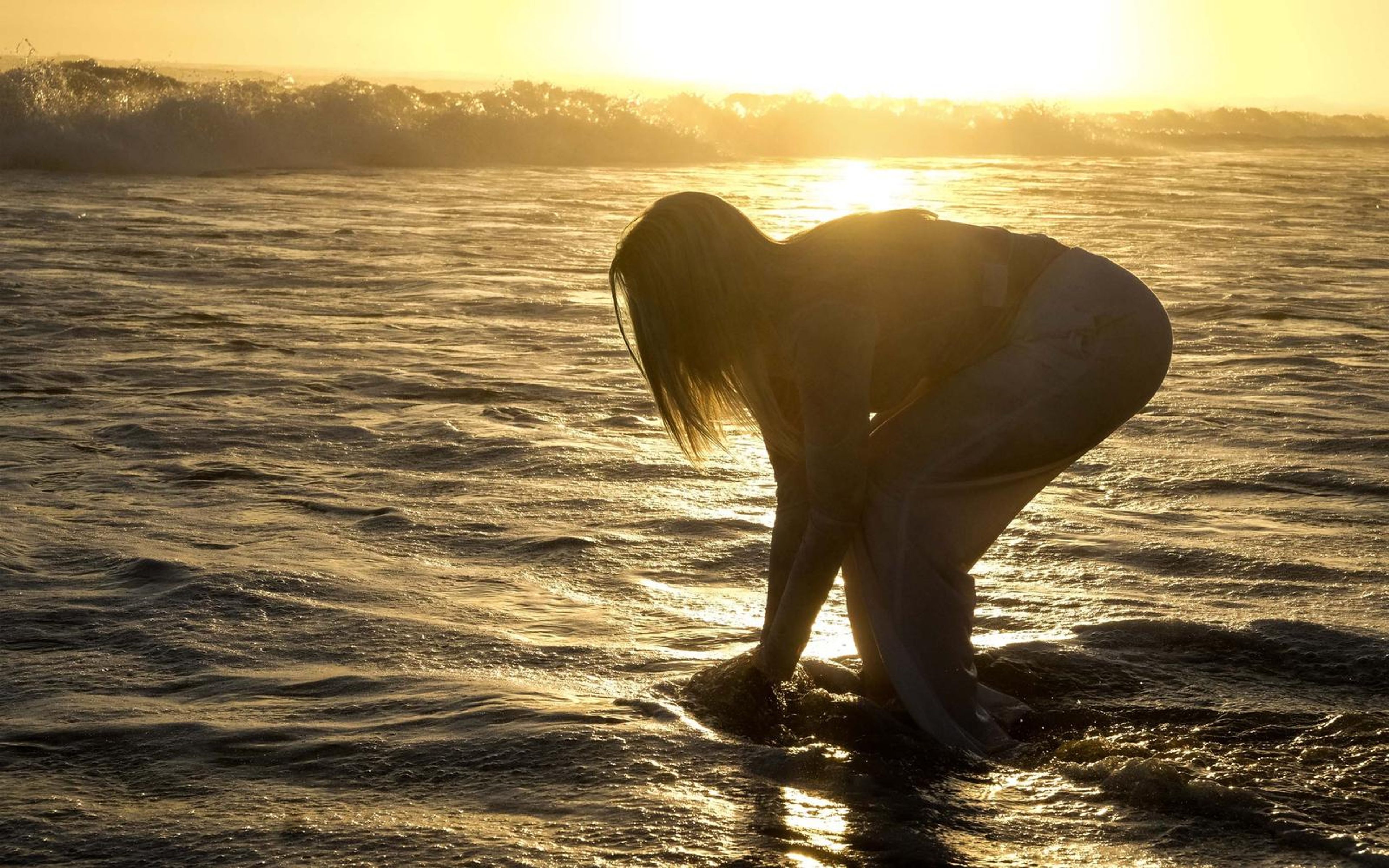 Woman bent forward touching the water in the surf, silhouetted by the low sun