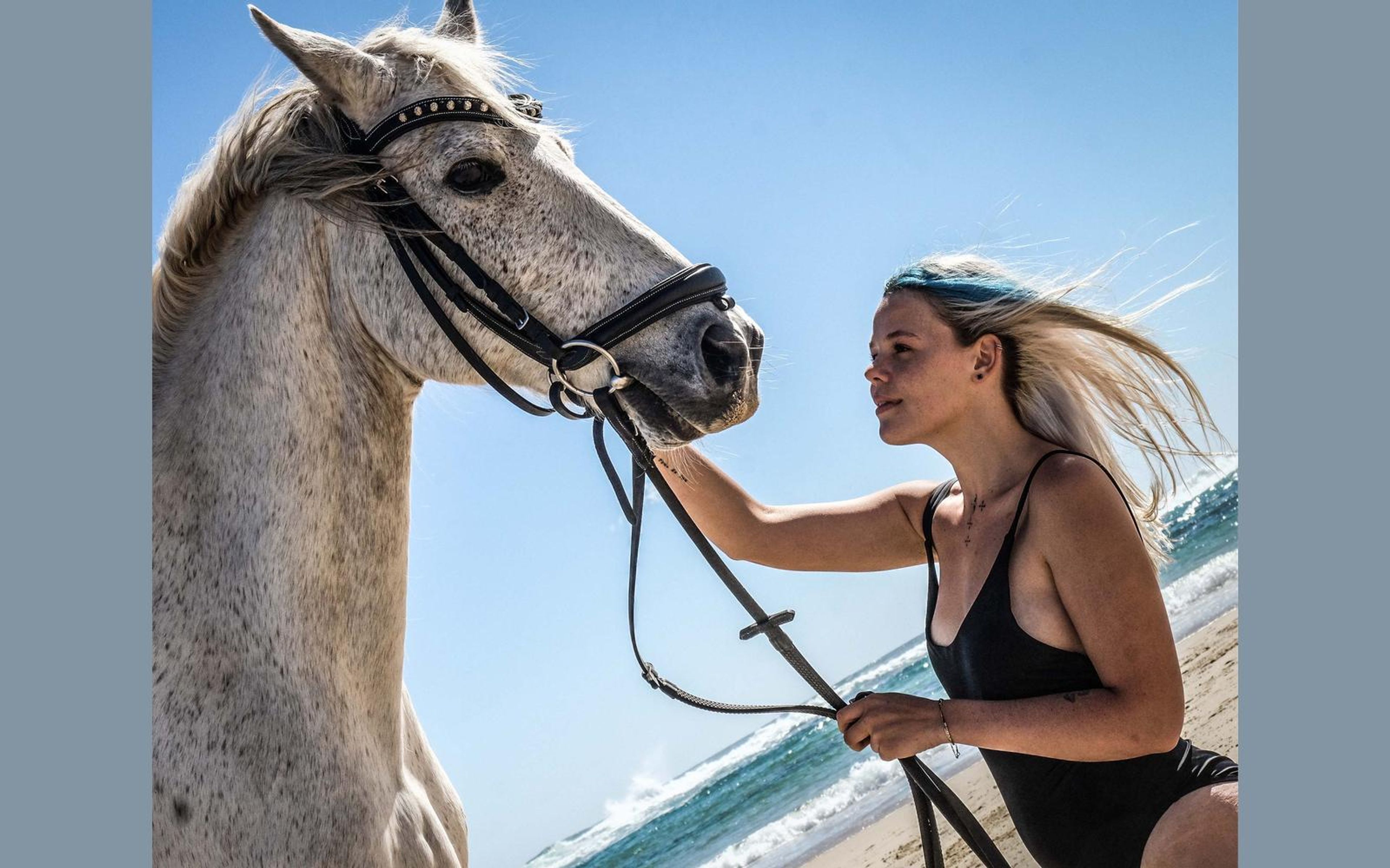Woman in a black swimsuit holding the reins beside a horse with the ocean behind her