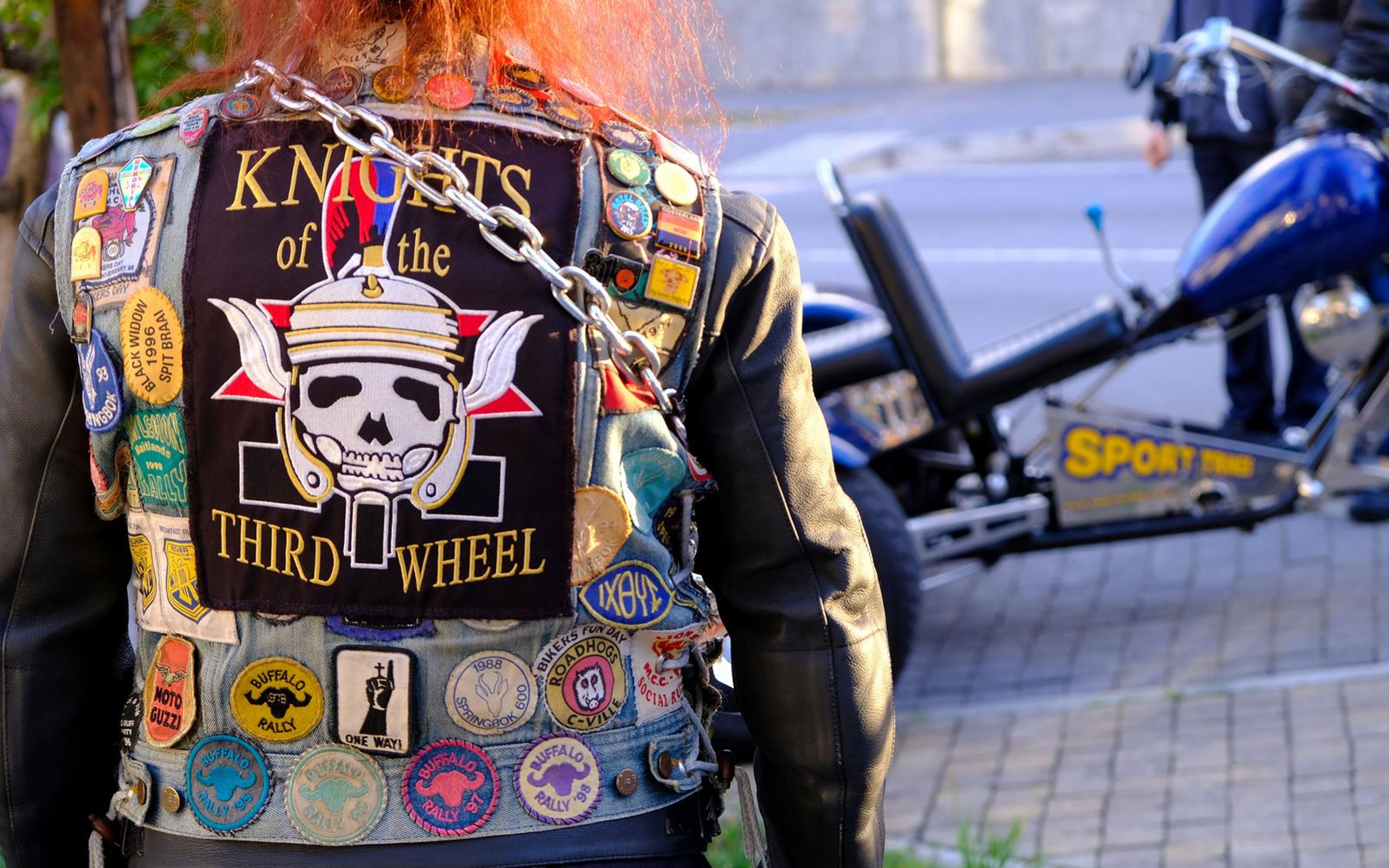 Back of a patched denim vest with a large skull-and-helmet club patch, rally badges and a heavy chain