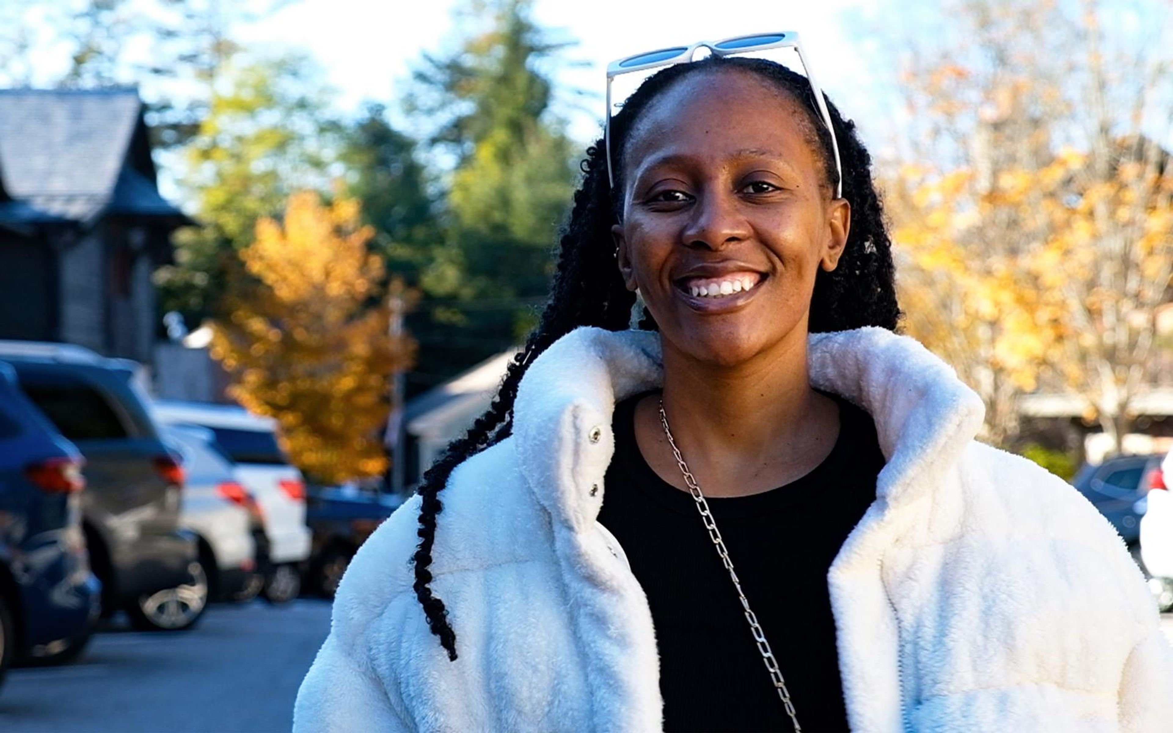 Model smiling at camera with white sunglasses pushed up on her head, long braids, white fur jacket