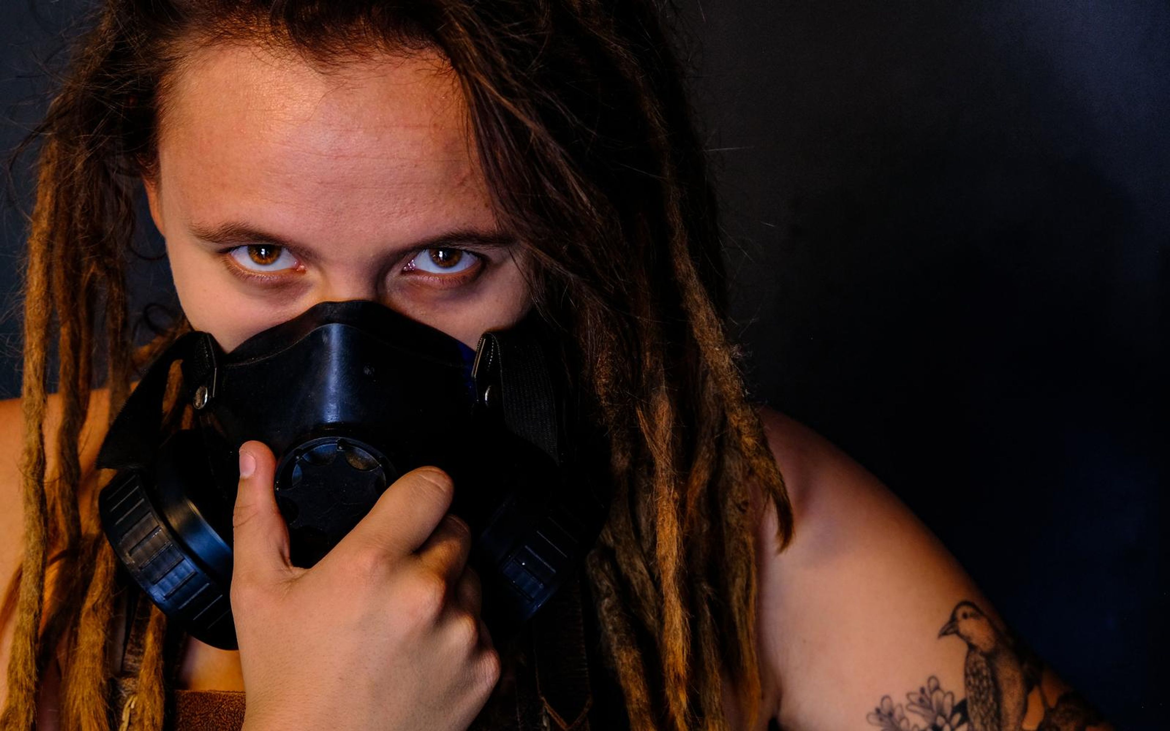 Close portrait of a woman peering over a black respirator mask with a hand at her chin, warm colour