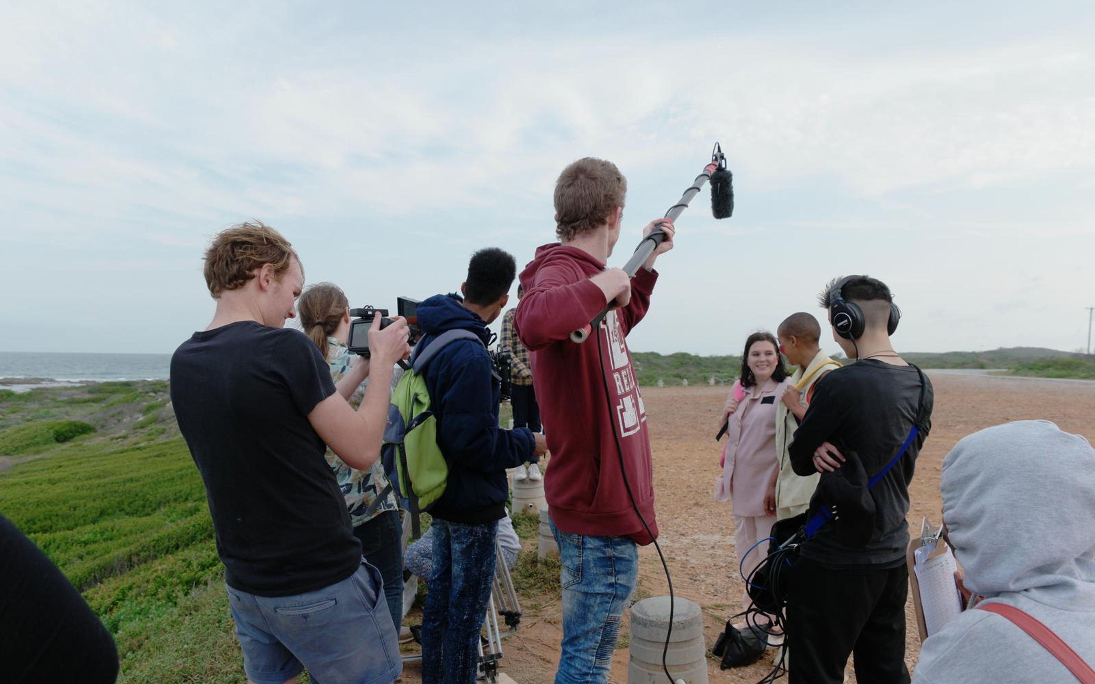 Crew gathered on a coastal lot with a boom pole raised over two actors in pastel uniforms